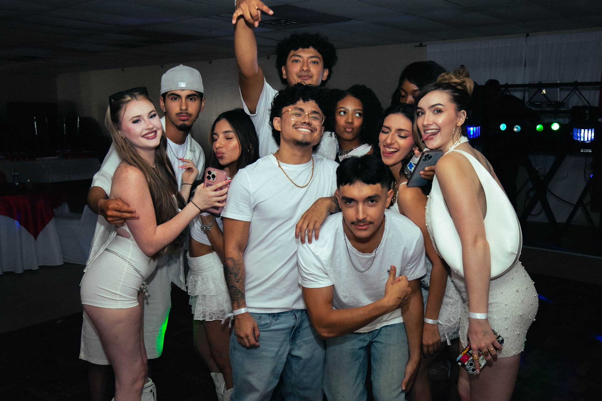 Group of young adults at a party posing for a group photo, some holding phones, smiling and making gestures.