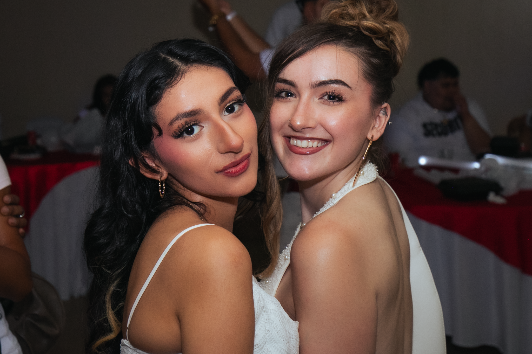 Two young women smiling and hugging at a social event, one with dark hair and the other with light hair in an updo, both wearing white dresses.