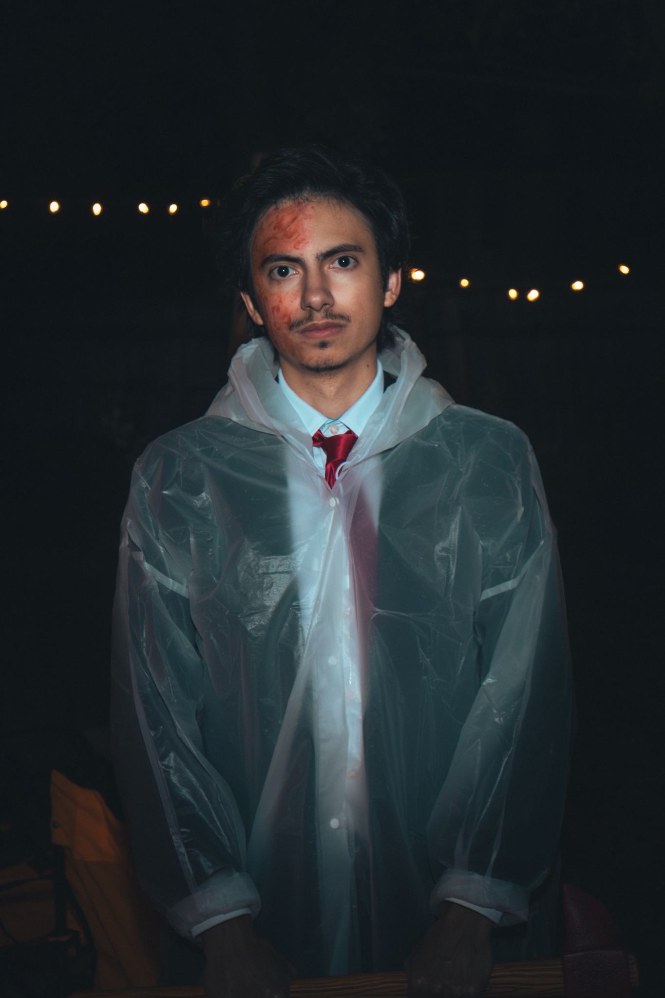 A man dressed in formal attire with a red tie and white shirt, wearing a clear plastic raincoat, stands outdoors at night with string lights in the background. He has dark hair and unusual red marks on his face.