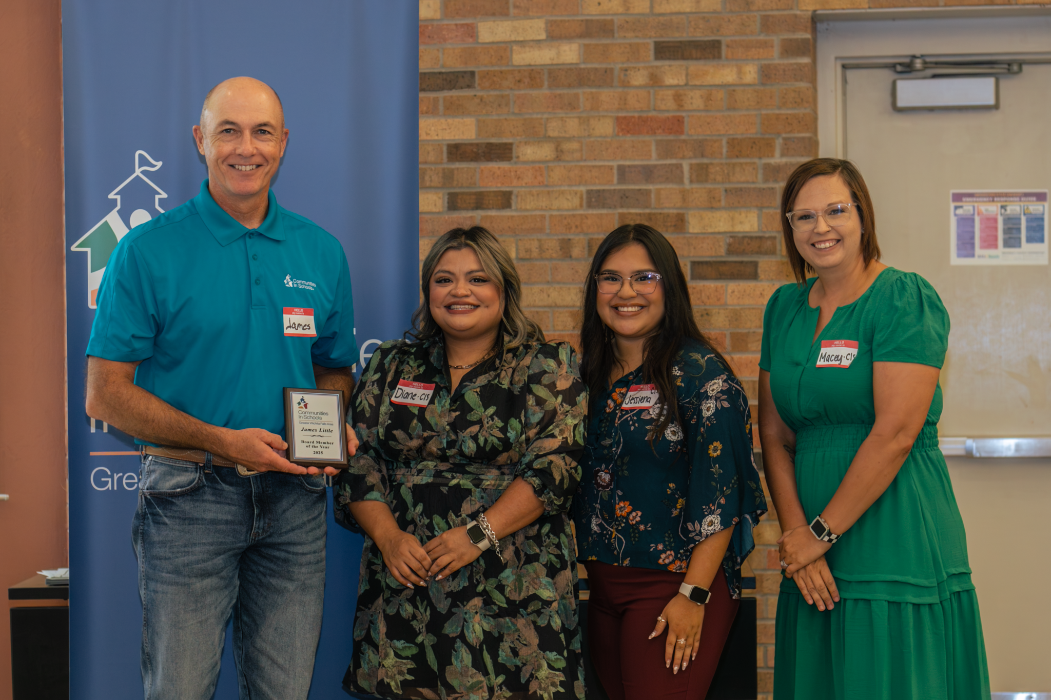 Four people standing indoors, three women and one man, smiling for a photo during an award or recognition event. The man is holding a plaque, and all are wearing name tags. Background includes a blue banner, a brick wall, and a door.