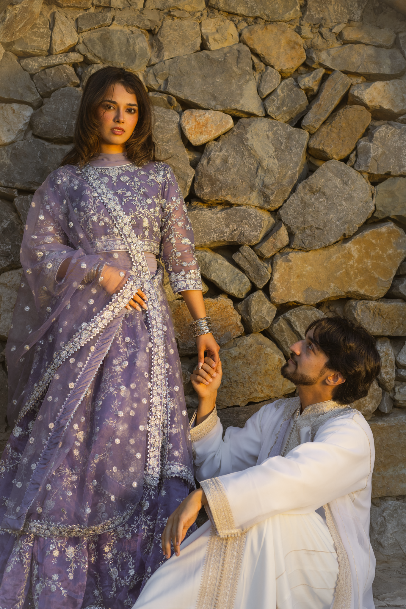 A man and woman dressed in traditional South Asian attire; the woman wears a purple embroidered outfit and the man wears a white kurta with gold embroidery; they hold hands against a stone wall, with the man sitting and looking up at the woman.