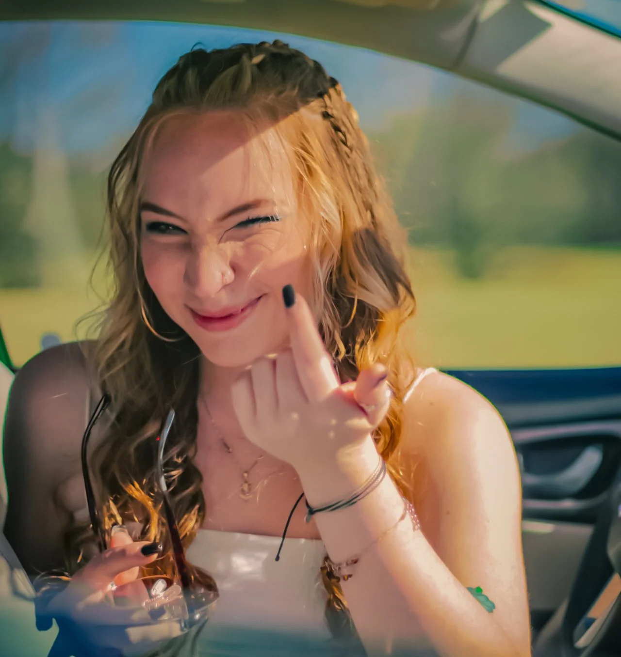 A young woman with curly red hair and a braid, smiling and making a playful gesture with her finger, sitting inside a vehicle with a blurred outdoor background.
