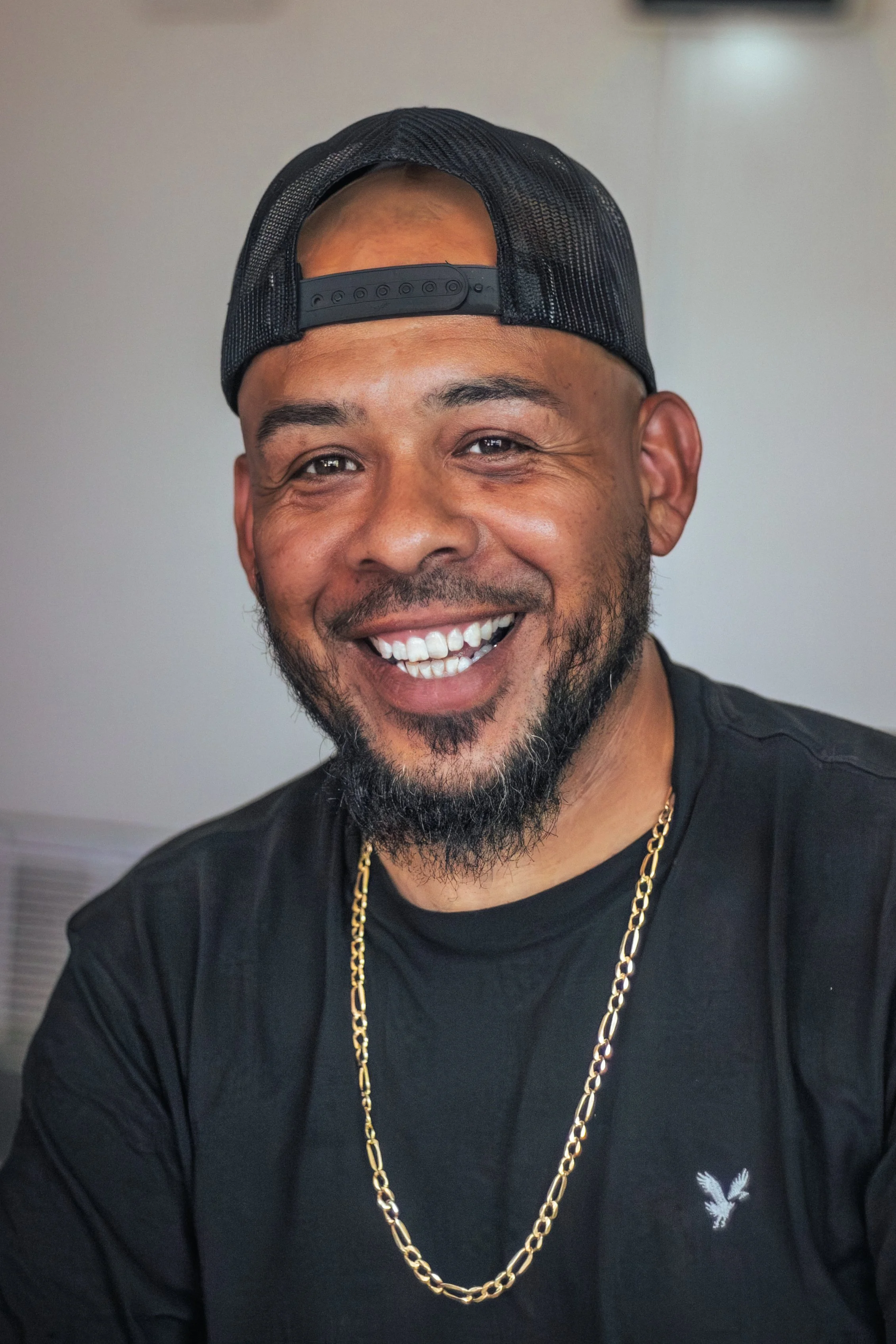 A smiling man wearing a black baseball cap backwards, a gold chain, and a black shirt with a small eagle logo, in front of a plain light-colored background.