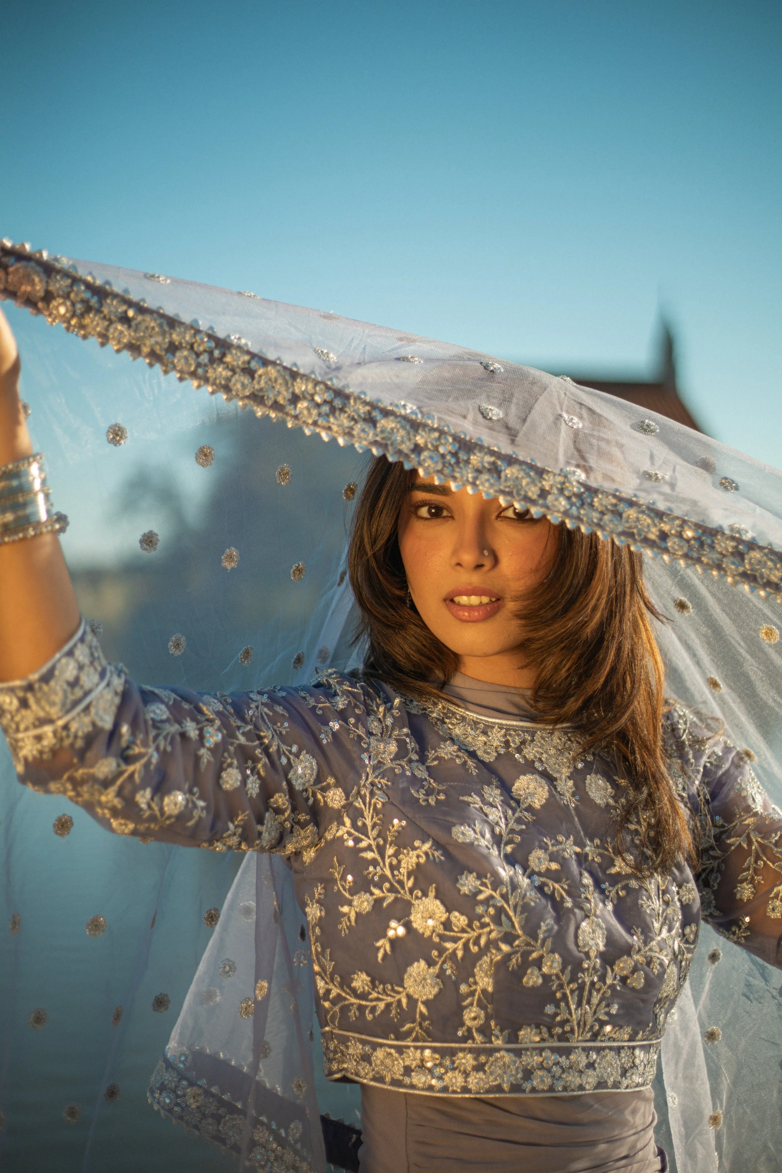 A young woman wearing traditional ethnic clothing and jewelry, holding a transparent parasol with gold embellishments, outdoors in front of a body of water during the day.