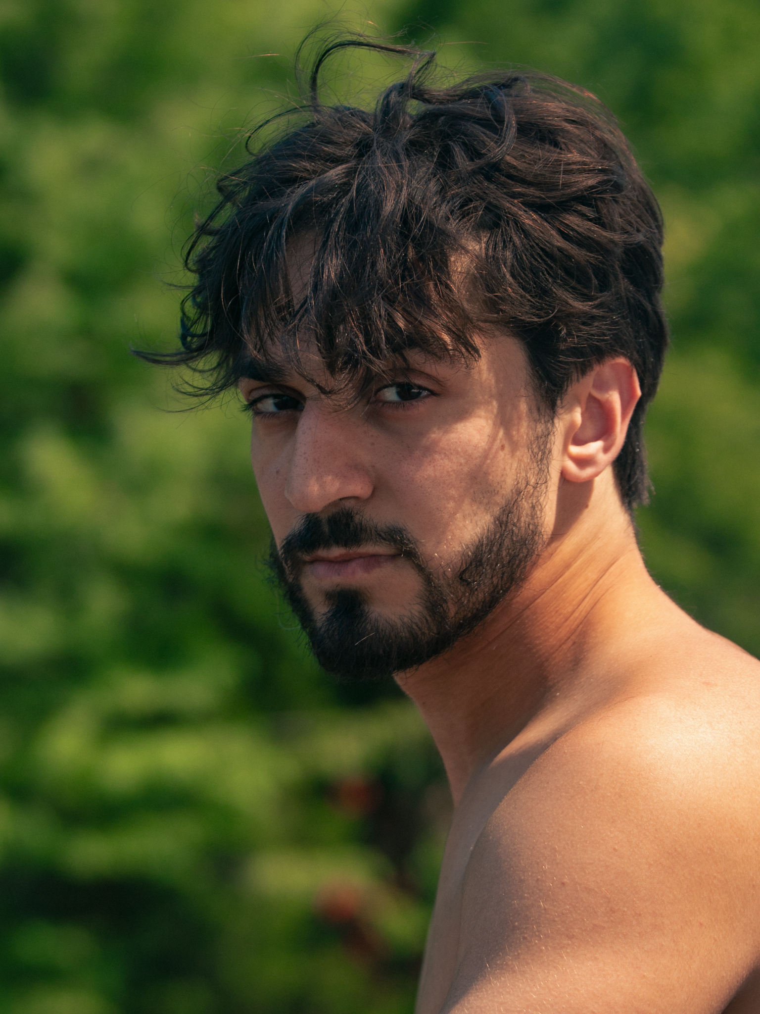 Close-up of a shirtless man with dark wavy hair and a beard, outdoors with green foliage in the background.