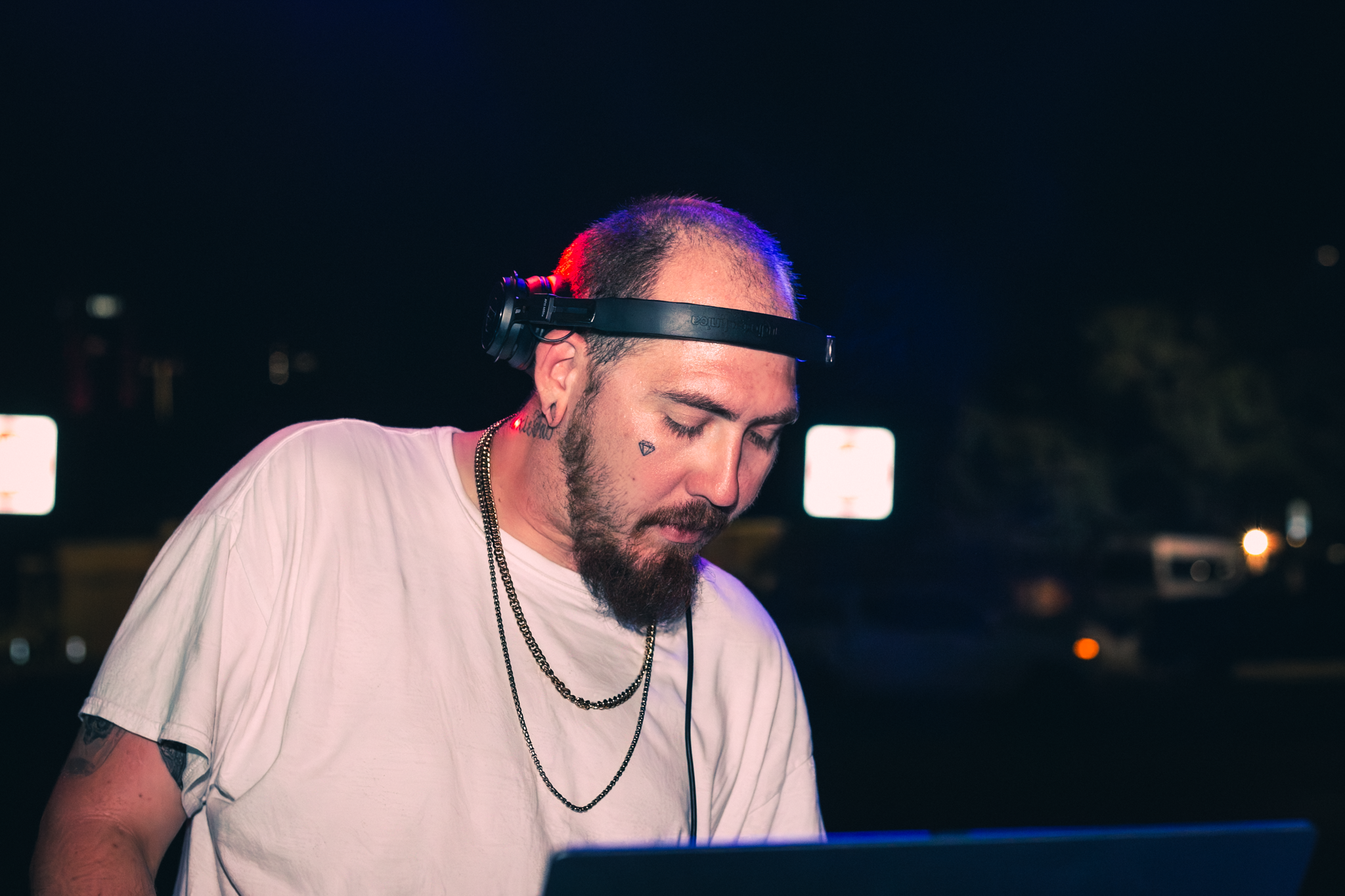 A DJ with short hair, facial tattoos, and a beard is mixing music at night, wearing headphones and a white t-shirt.