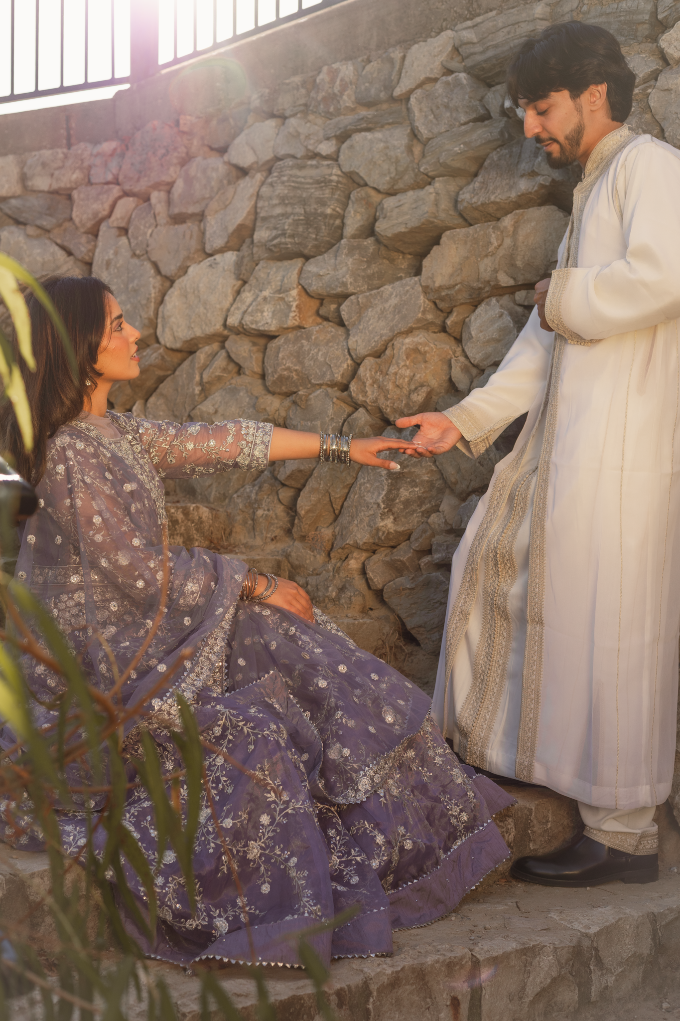 A man dressed in traditional South Asian attire offers a coin to a seated woman wearing a purple embroidered dress, in front of a stone wall with sunlight shining from behind.