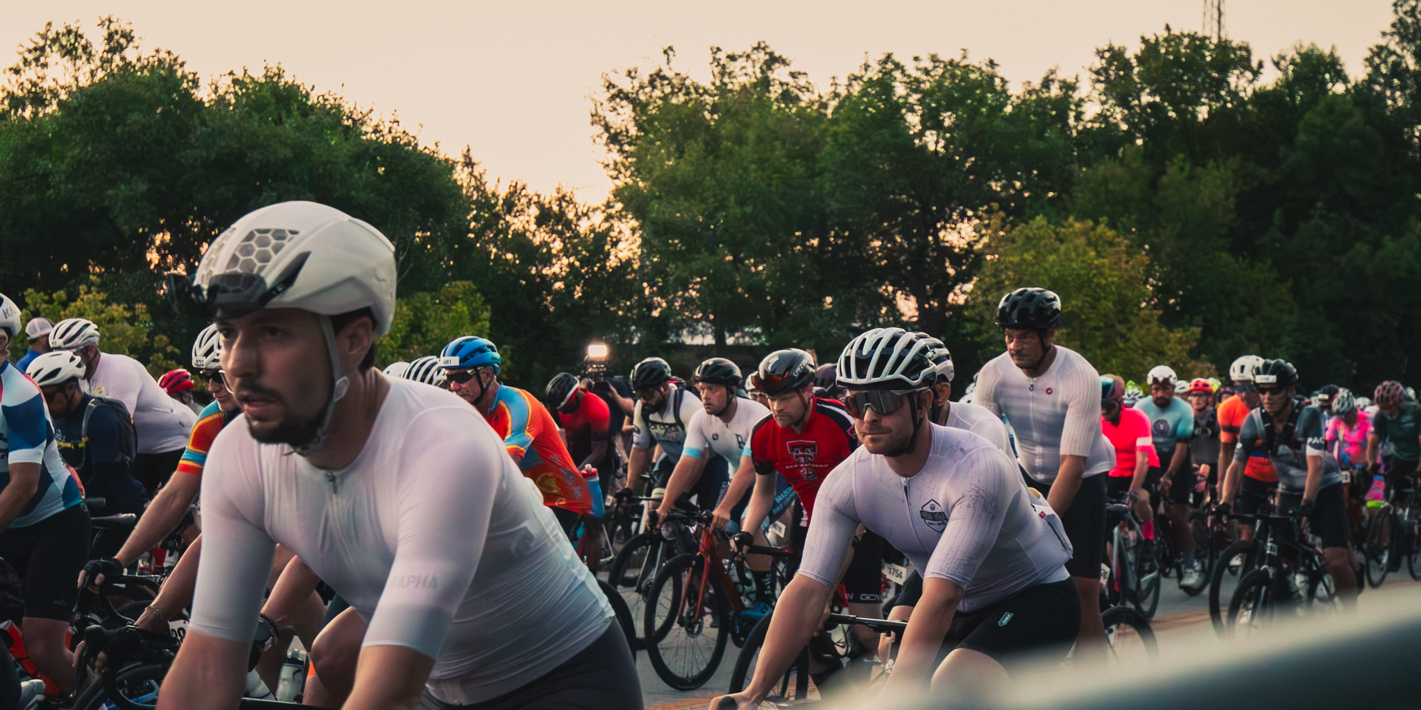 A large group of cyclists wearing helmets and athletic clothing participating in a bike race or event outdoors during sunset, with trees in the background.