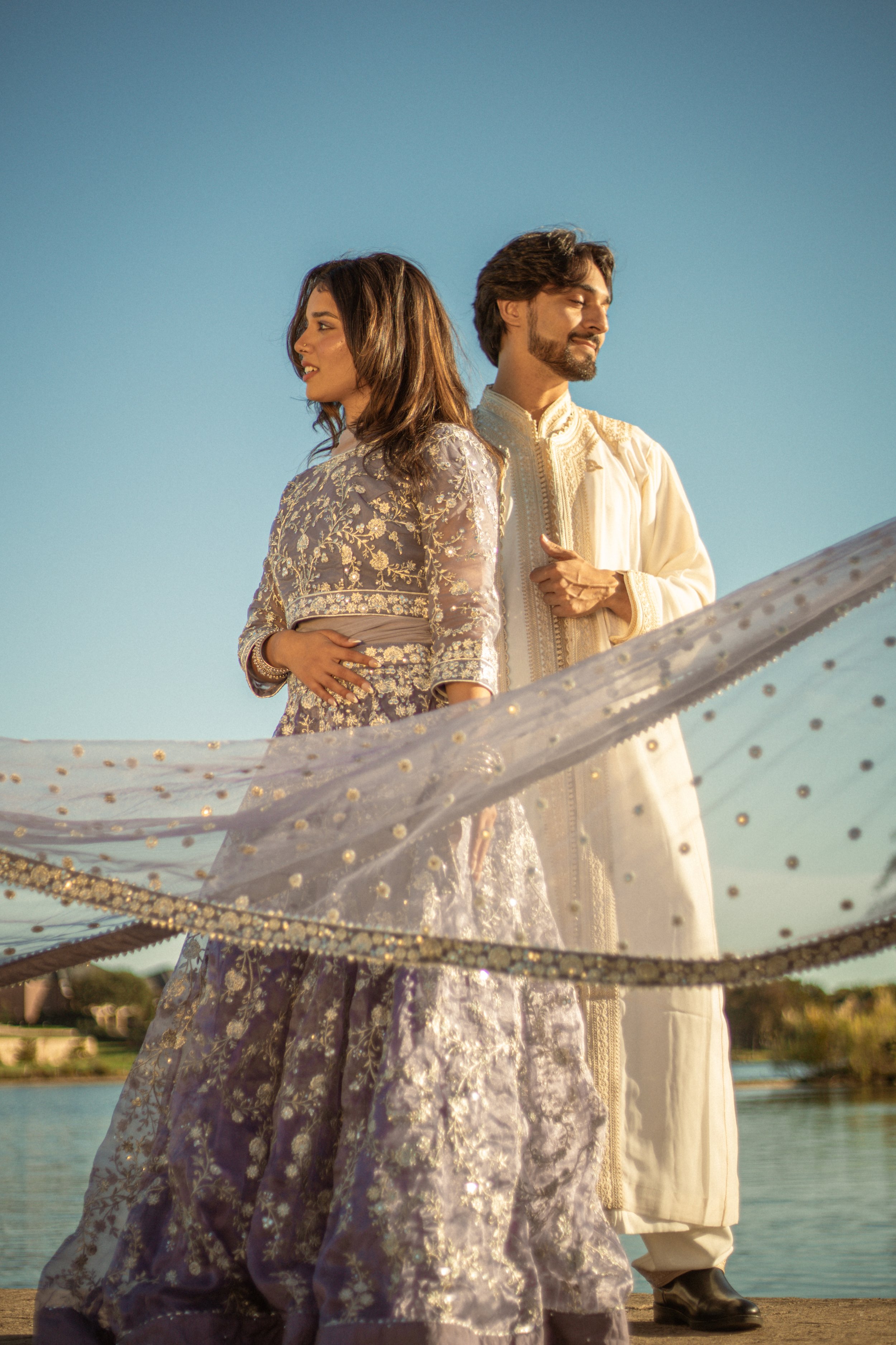 A man and woman standing back-to-back outdoors near a body of water, both dressed in traditional South Asian attire with intricate embroidery. The woman is wearing a purple and gold lehenga, and the man is wearing an off-white kurta with embroidery. 