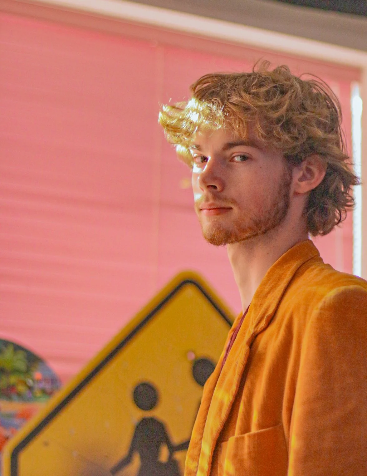 A young man with wavy blonde hair and a light beard wearing an orange jacket. He is looking at the camera with a slight smile, standing in front of a pink wall with a street sign and colorful artwork.