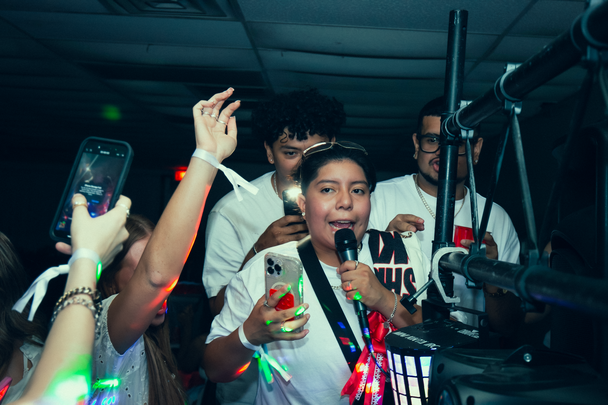 A group of young people celebrates at a party, with some holding phones and others standing around a woman speaking into a microphone. Colorful lights are visible, and the scene appears lively and festive.