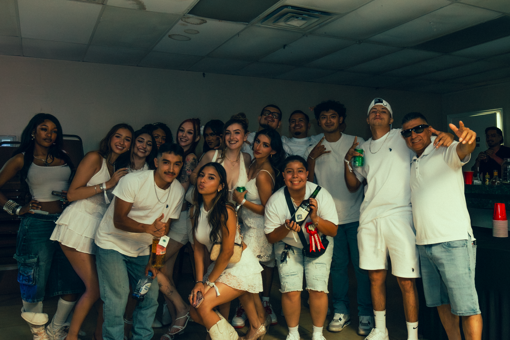 Group of young adults and adults posing together at a party, some holding drinks, inside a room with a low ceiling and a bar in the background.