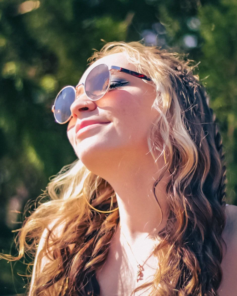 Young woman with wavy hair wearing sunglasses outdoors, smiling with her eyes closed, enjoying sunlight.
