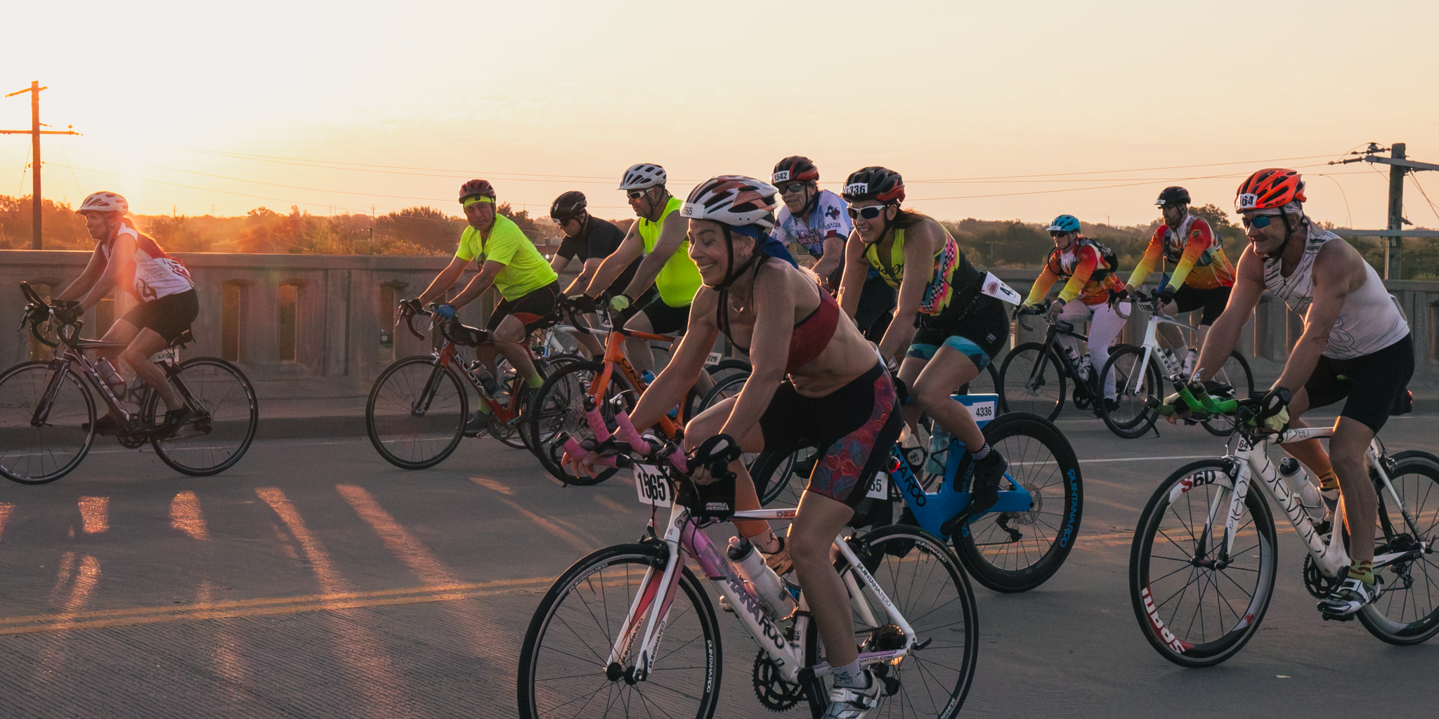 A group of cyclists participating in a race during sunset, riding on a bridge with utility poles and trees in the background.