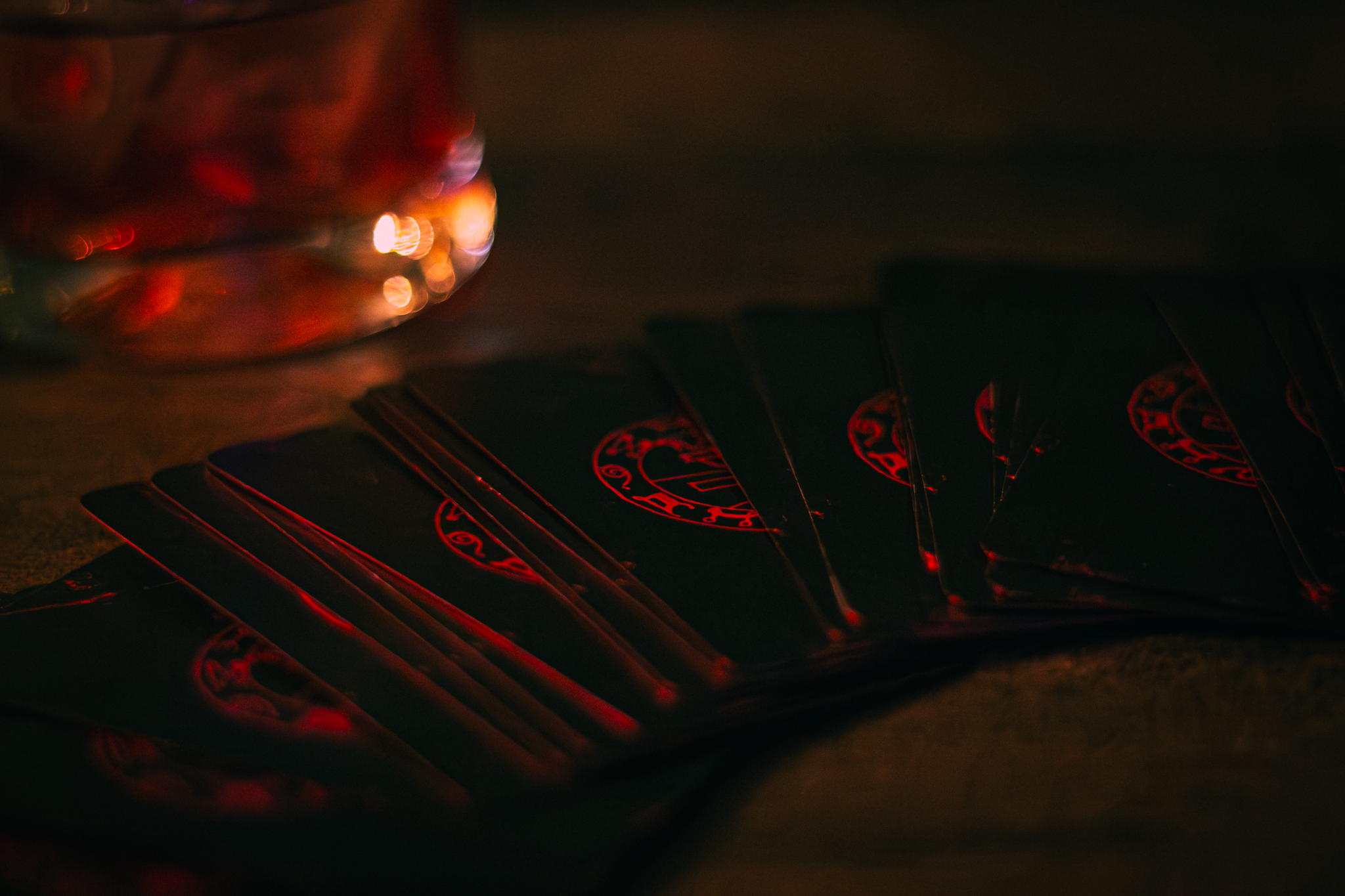 A row of playing cards with red and black backs laid out on a dark surface, with a glass of a dark beverage in the background, illuminated by dim red lighting.