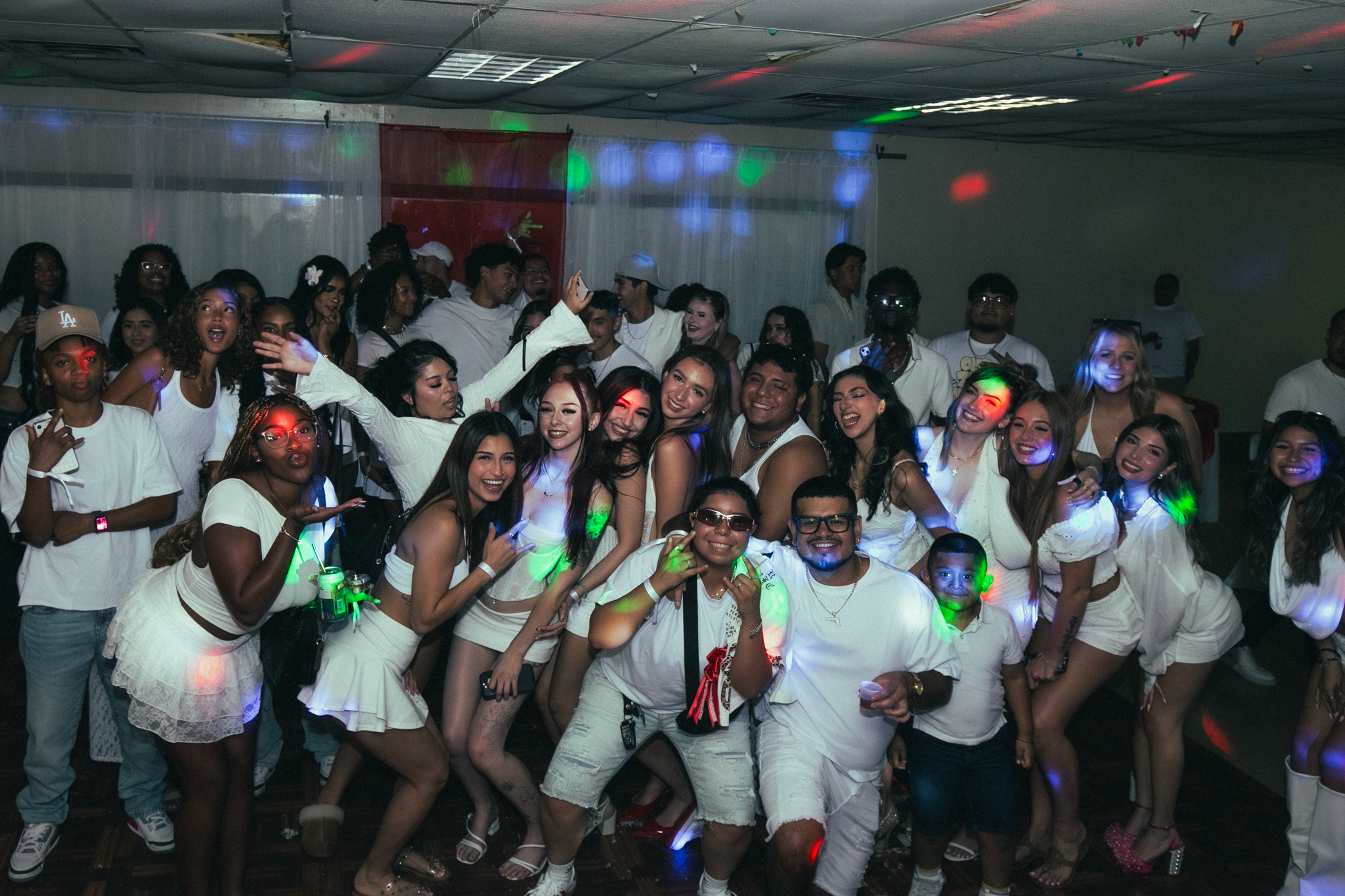 A group of people at a party, dressed in white, dancing and smiling in a dimly lit room with colorful lights.
