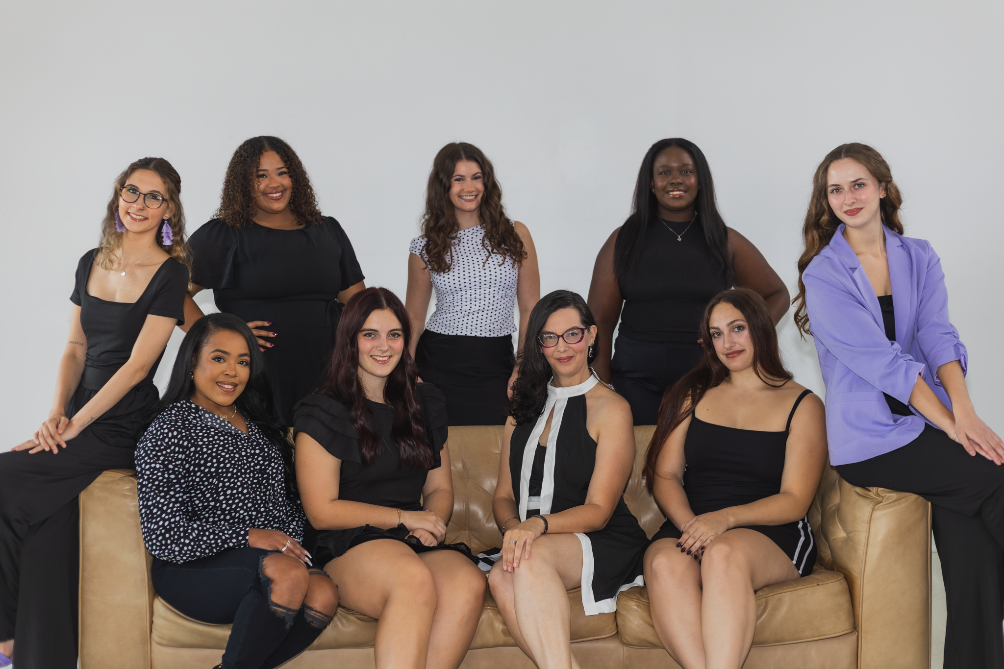 A group of eleven diverse women posing together in a studio with a plain white background. Some are seated on a tan leather couch, others standing behind or sitting on the armrest, dressed in black, white, and purple outfits, smiling at the camera.