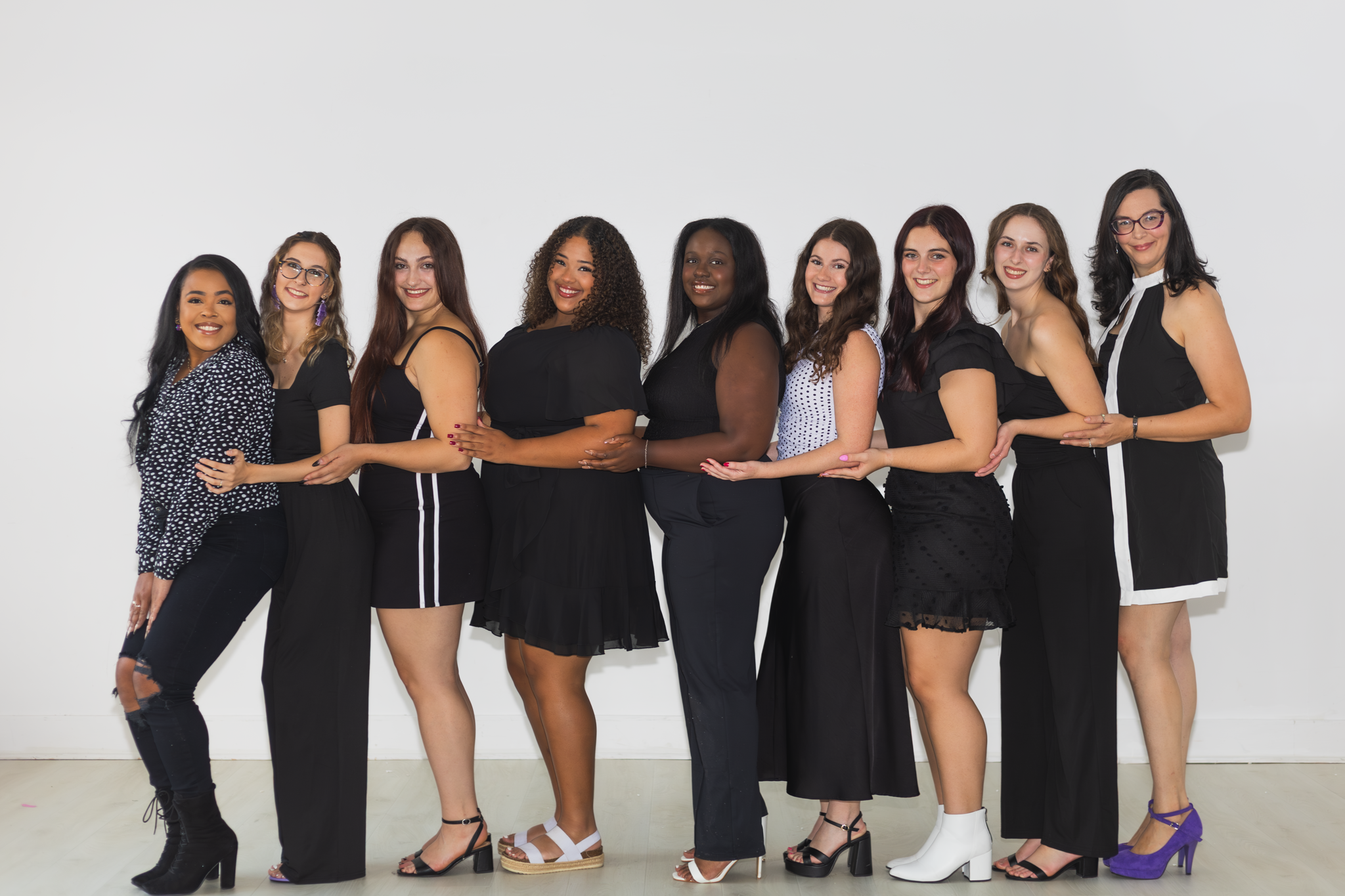 Group of nine women standing in a line, holding each other by the waist or shoulders, smiling, against a plain white background.