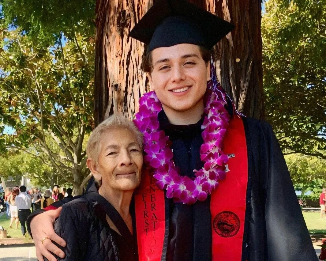 A young man in graduation cap and gown with a purple lei around his neck, standing beside an older woman outdoors, both smiling, with trees and people in the background.
