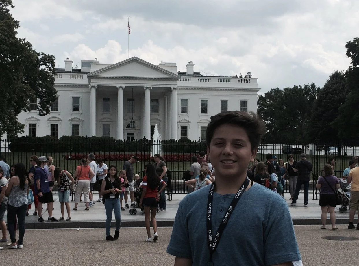 A young boy smiling in front of the White House during daytime with crowds of people gathered behind a black fence.