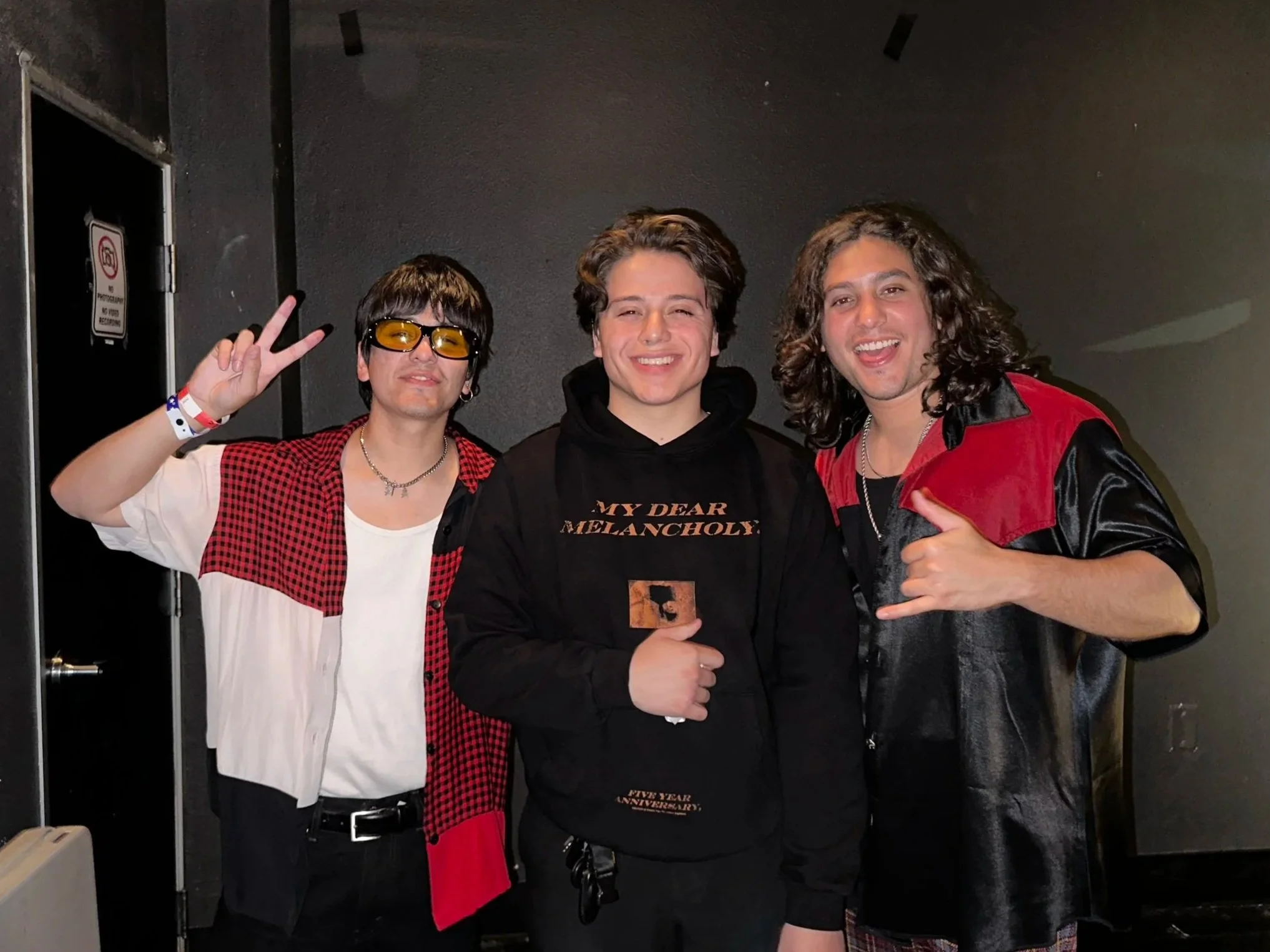 Three young men standing together, smiling, in a dark indoor setting. The person on the left is wearing yellow-tinted glasses, a white T-shirt, and a red plaid shirt. The middle person is dressed in a black hoodie with text, and the right person is wearing a black and red shiny jacket, with long curly hair. All are casual and appear happy.