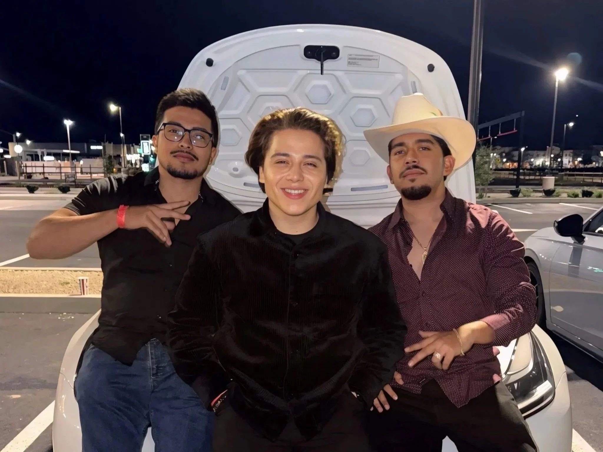 Three men standing in front of an open car hood at night in a parking lot, with streetlights and empty parking spaces in the background.