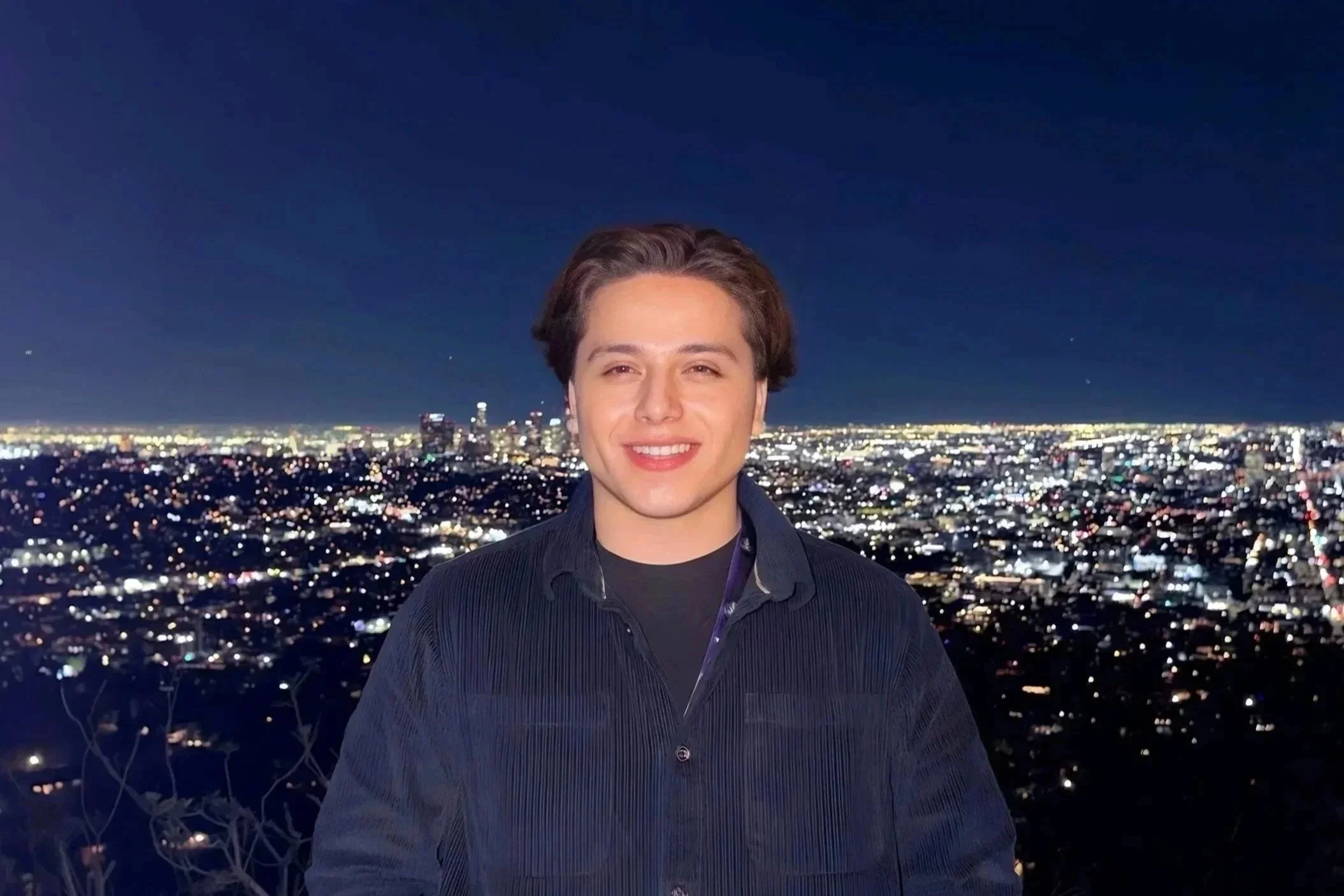 A young man with brown hair smiling at night, standing in front of a cityscape with numerous lights and tall buildings, under a dark sky.