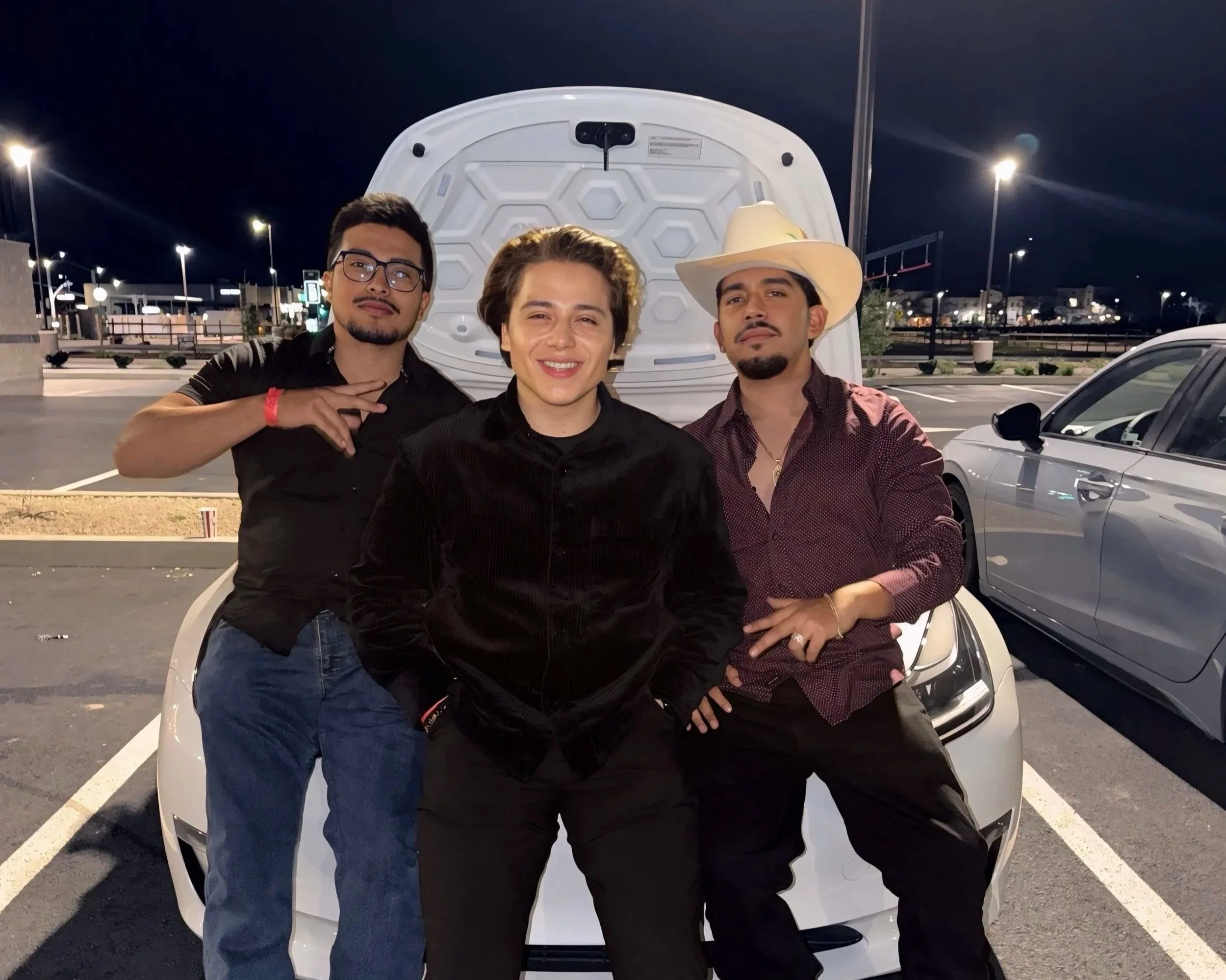 Three men standing together in front of a white car with its hood open at night in a parking lot, with city lights and streetlights visible in the background.
