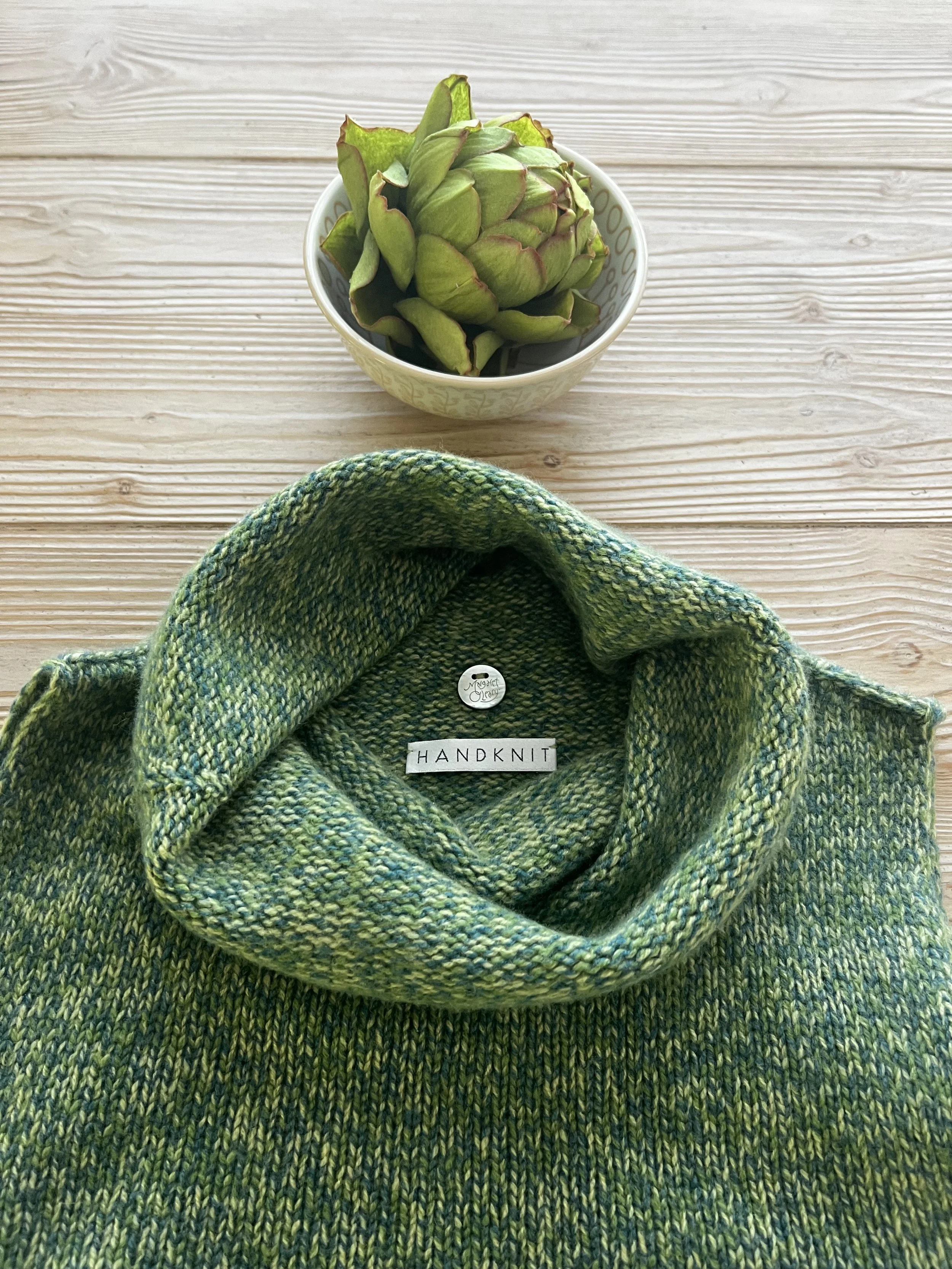 A green and yellow handknit scarf with a label that says "HANDKNIT" is draped on a light wooden surface with a single small plate of an artichoke in a bowl positioned above it.