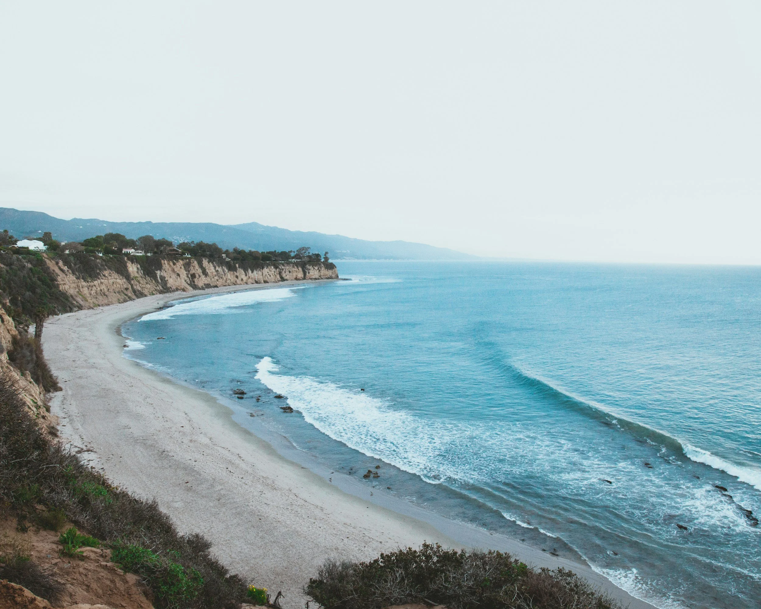 A scenic view of a beach with a curved shoreline, cliffs on the left side, and ocean waves, with mountains in the background under a light sky.