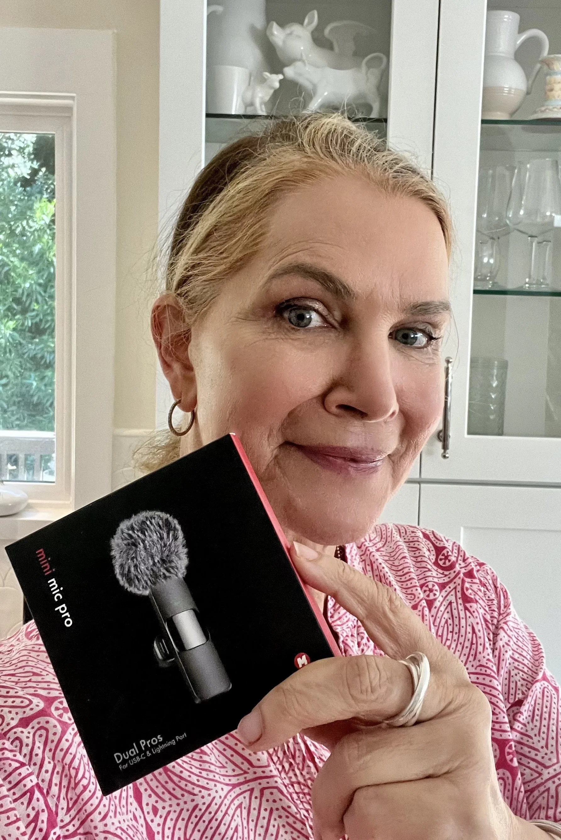 A woman with blonde hair, wearing a pink patterned shirt and hoop earrings, is holding a black microphone box in her left hand and standing in a kitchen near a glass-front cabinet with white ceramic animal figurines. The background shows a window with a green outdoor view.