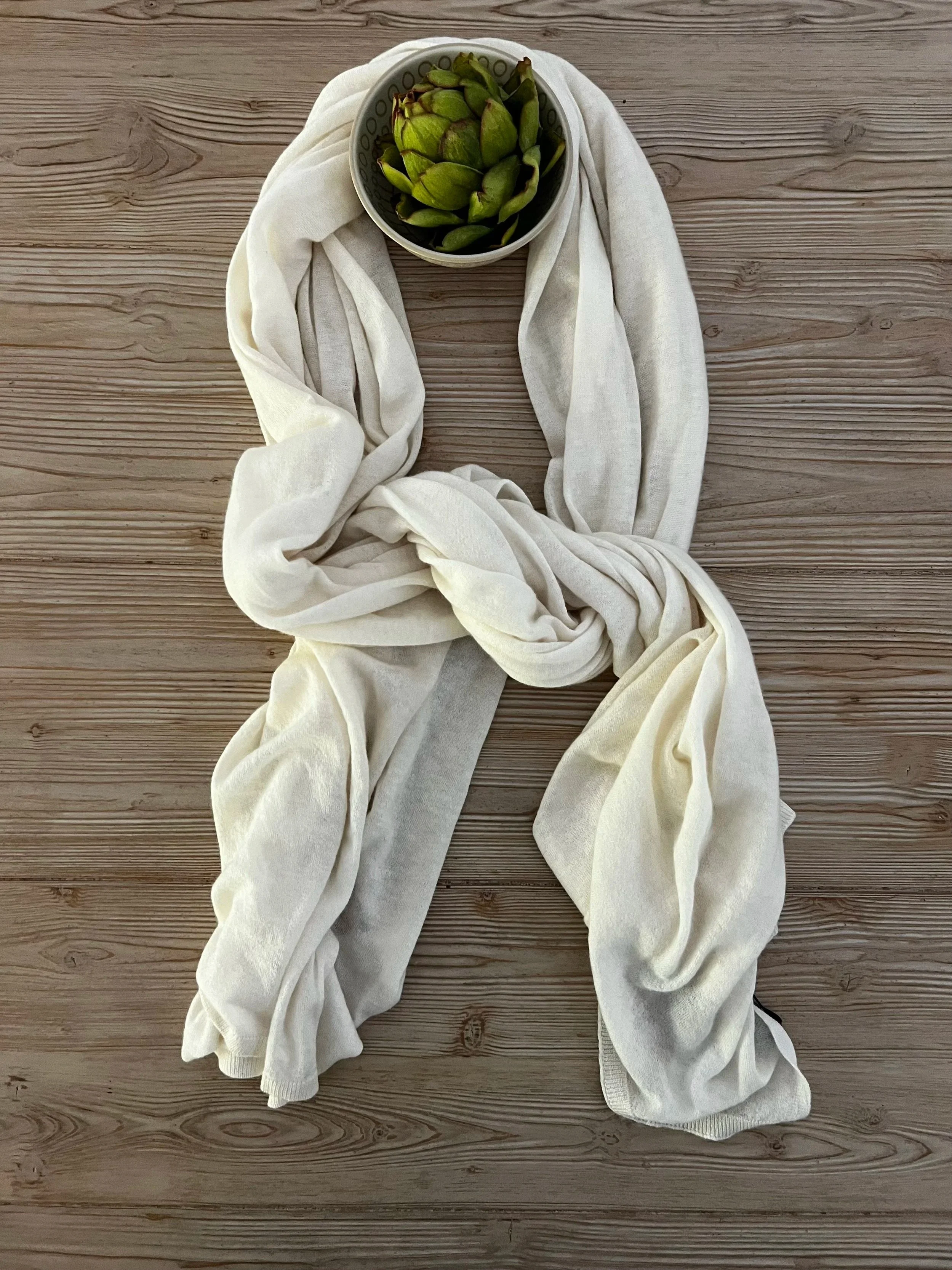 A lightweight cream cashmere scarf next to a bowl of green artichokes on a wooden surface.