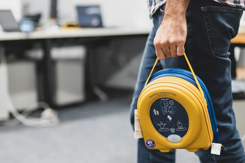 Person holding a yellow and blue Automated External Defibrillator (AED) with instructions on the front.