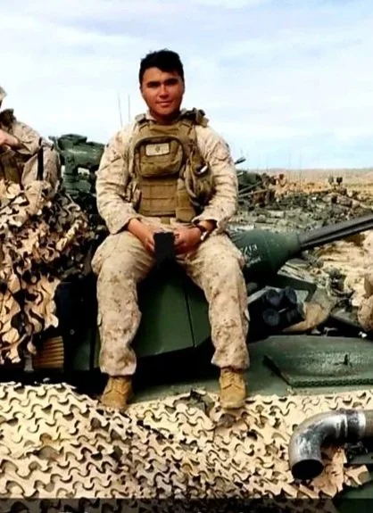 A soldier in camouflage uniform sitting on a military tank, outdoors in a desert environment.