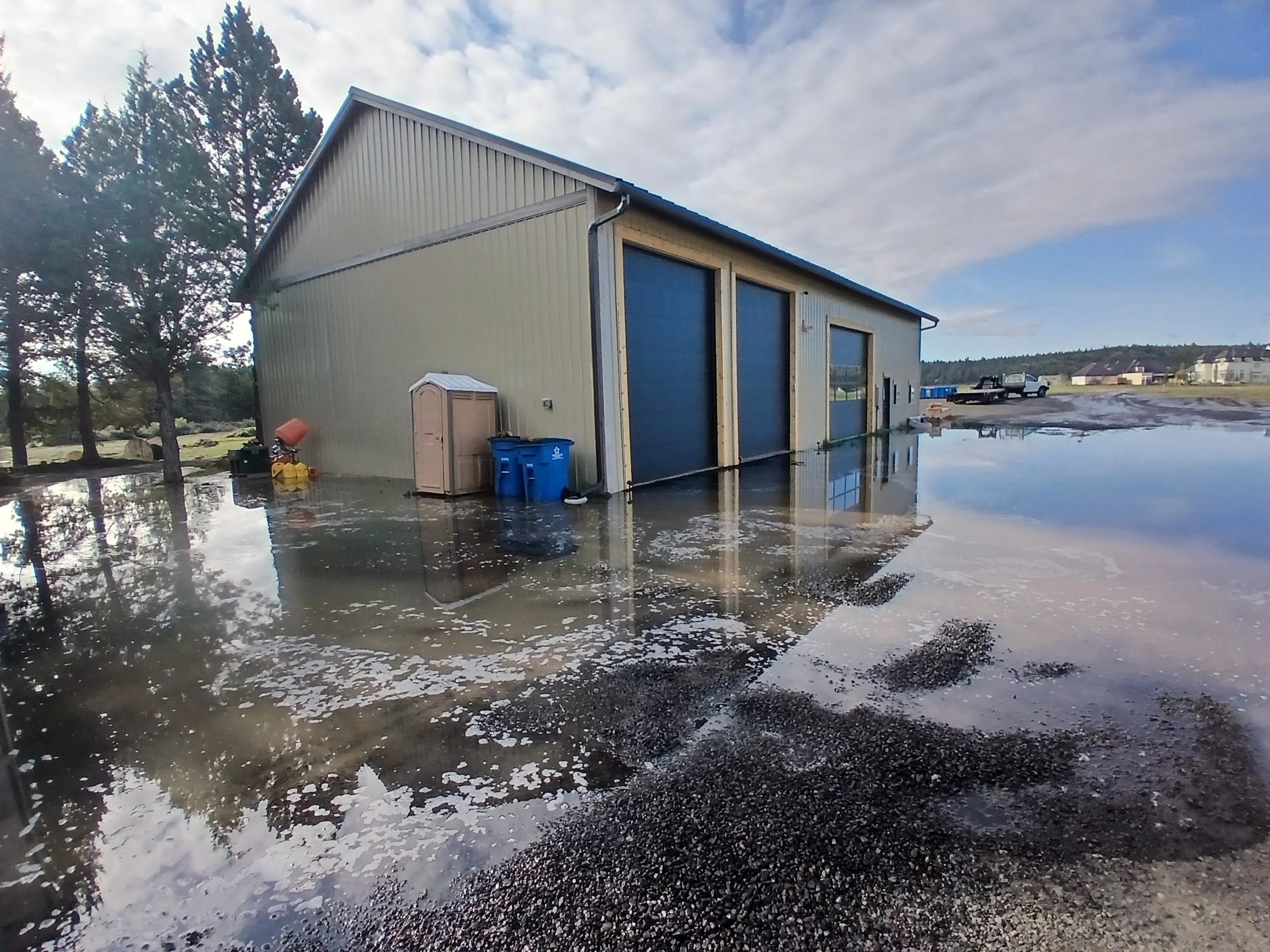 A large beige metal building partially submerged in floodwaters, with a small beige shed and blue recycling bins outside. Water surrounds the building, reflecting the sky and nearby trees. In the background, there are trees, houses, and a partly cloudy sky.
