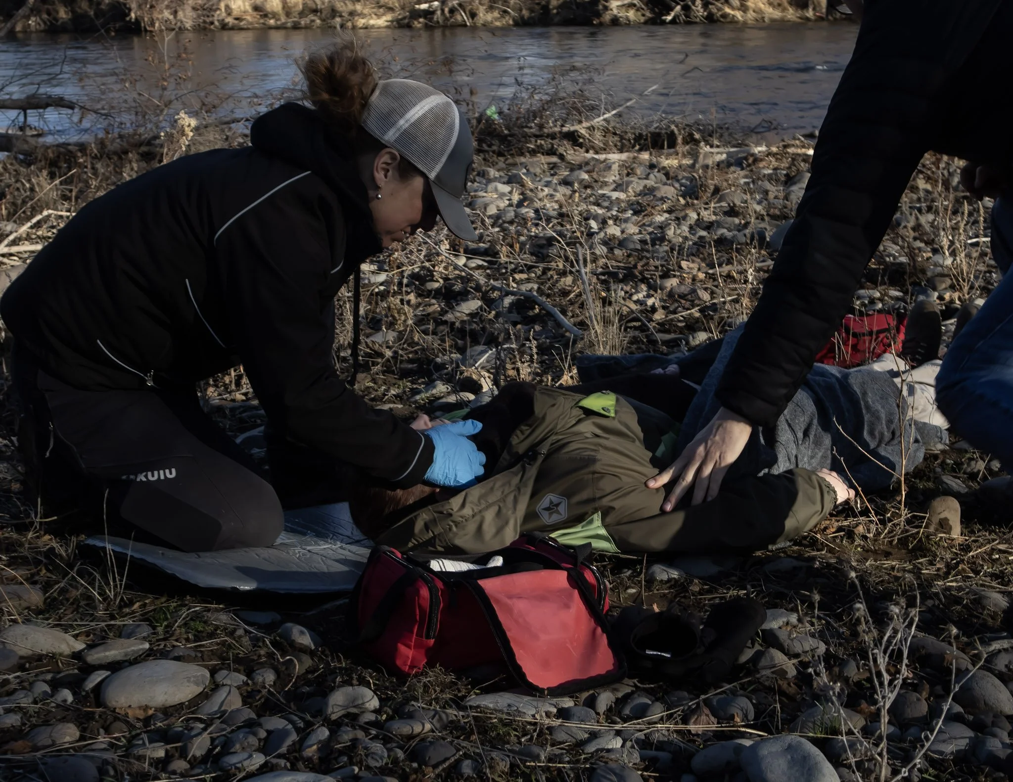 Two people providing medical assistance to a person lying on the ground near a riverbank, with emergency supplies nearby.