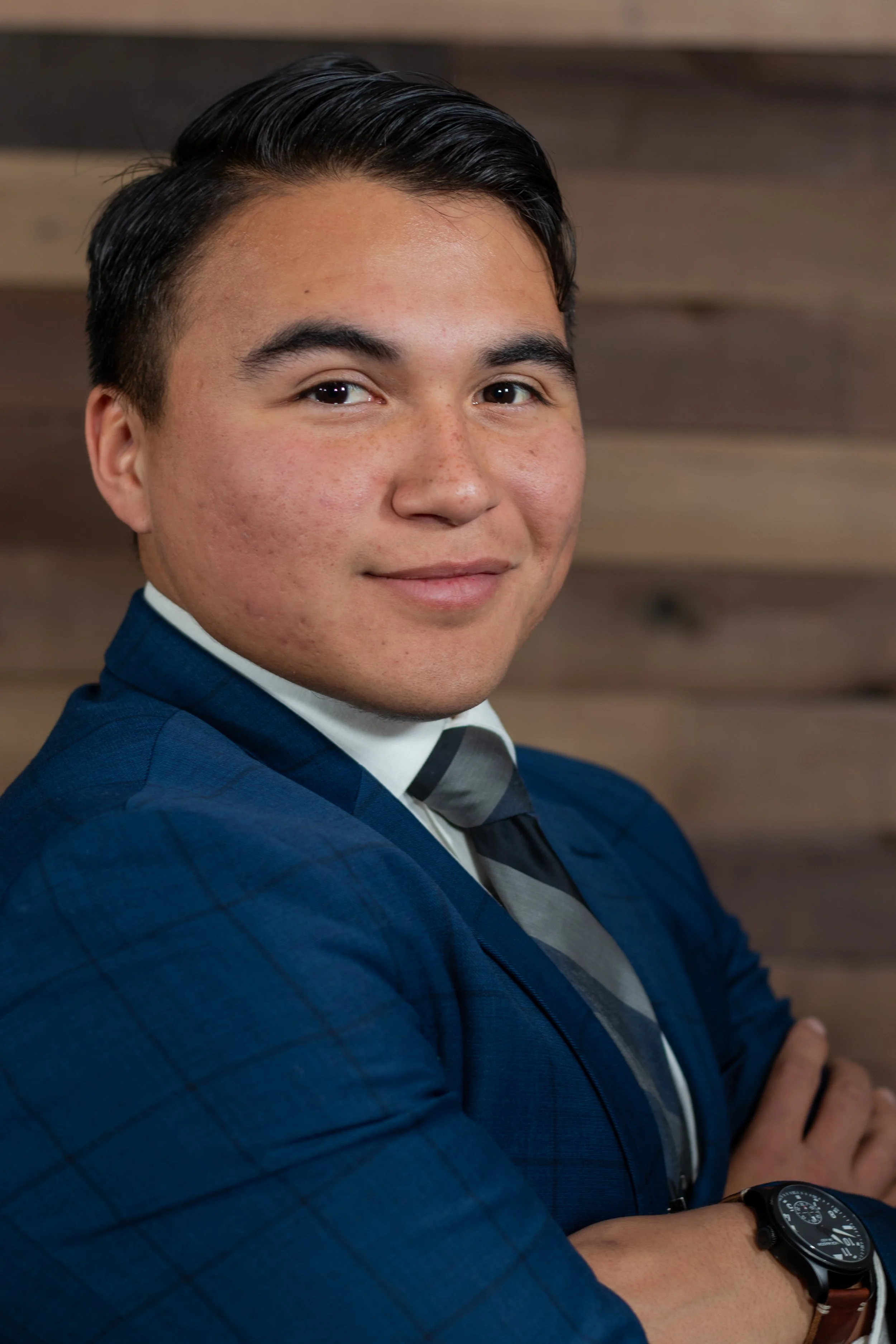 A young man with dark hair, wearing a blue checked suit, white shirt, and striped tie, standing with arms crossed against a wooden wall background.