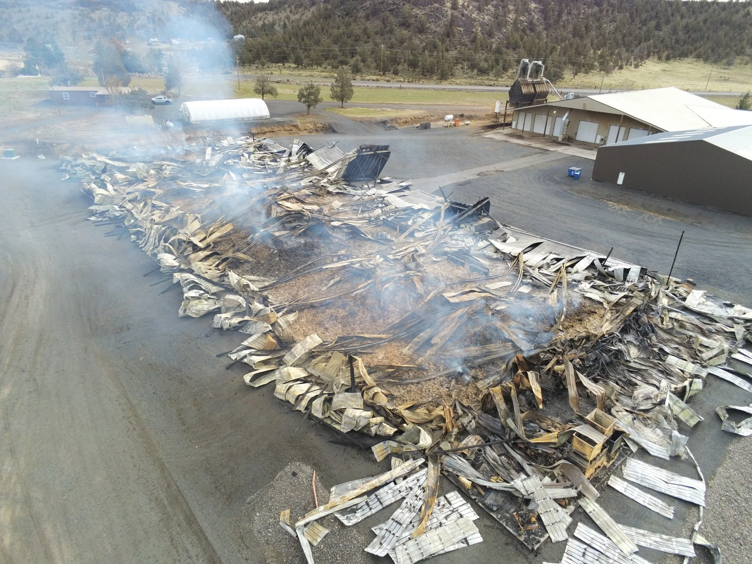Burned debris and wreckage from a collapsed metal building with smoke rising, located in an outdoor area with nearby structures and a gravel driveway.