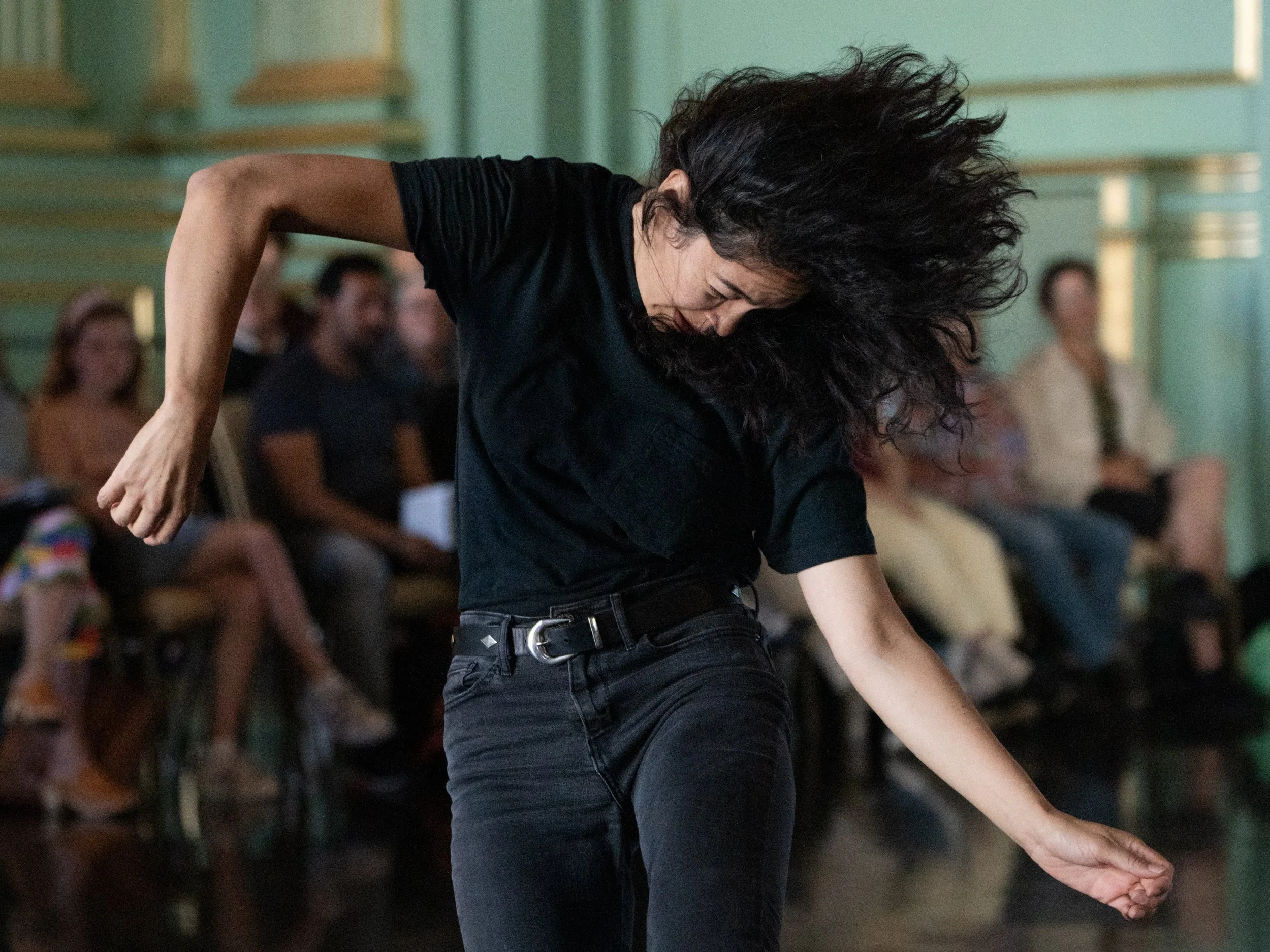 Woman with curly black hair dancing or performing in front of an audience in a room with green walls.