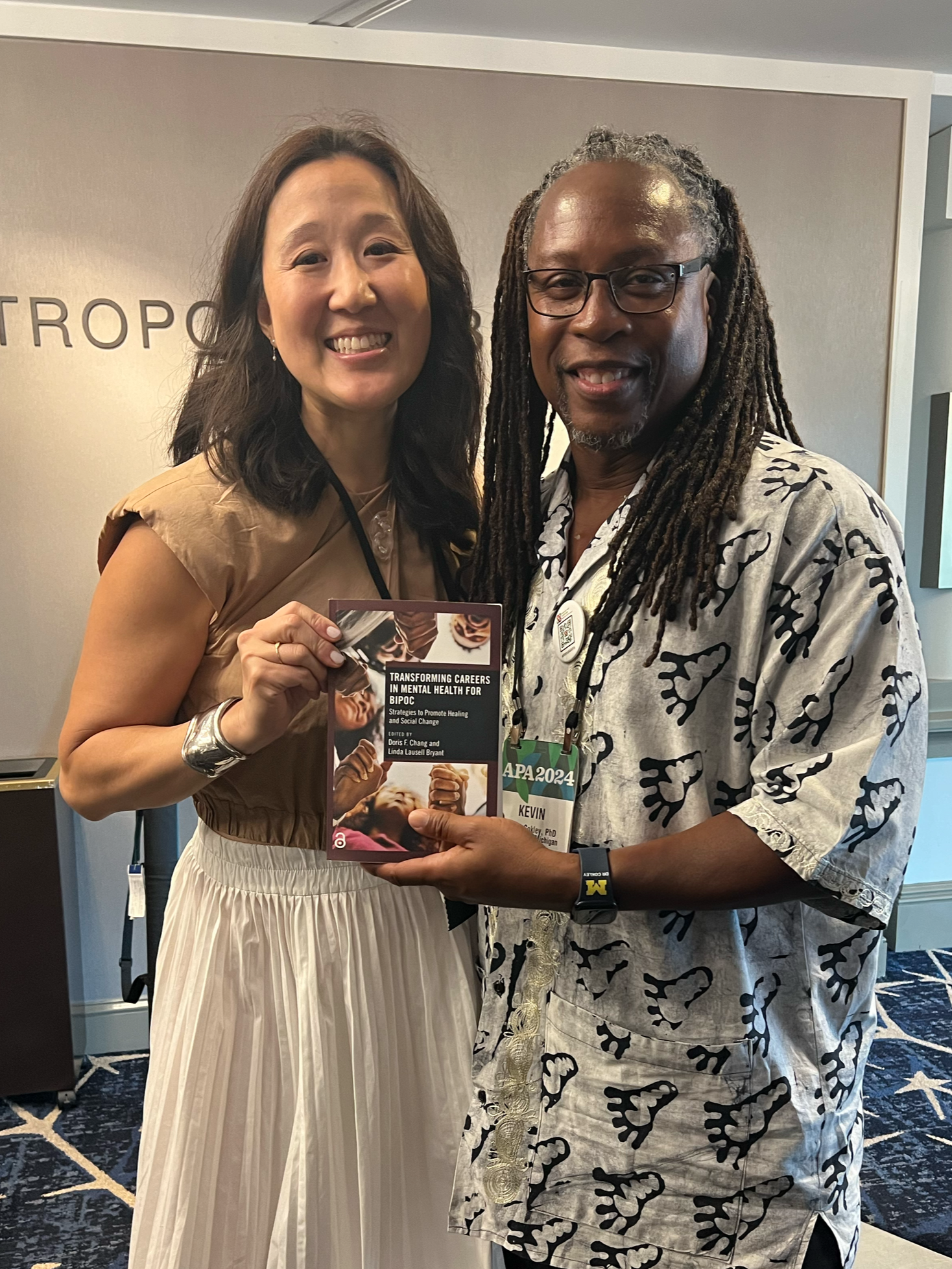 Two people smiling and holding a book at a conference. The woman on the left has shoulder-length dark hair and is wearing a beige top and cream-colored skirt. The man on the right has long dreadlocks, glasses, a patterned short-sleeve shirt, and is w