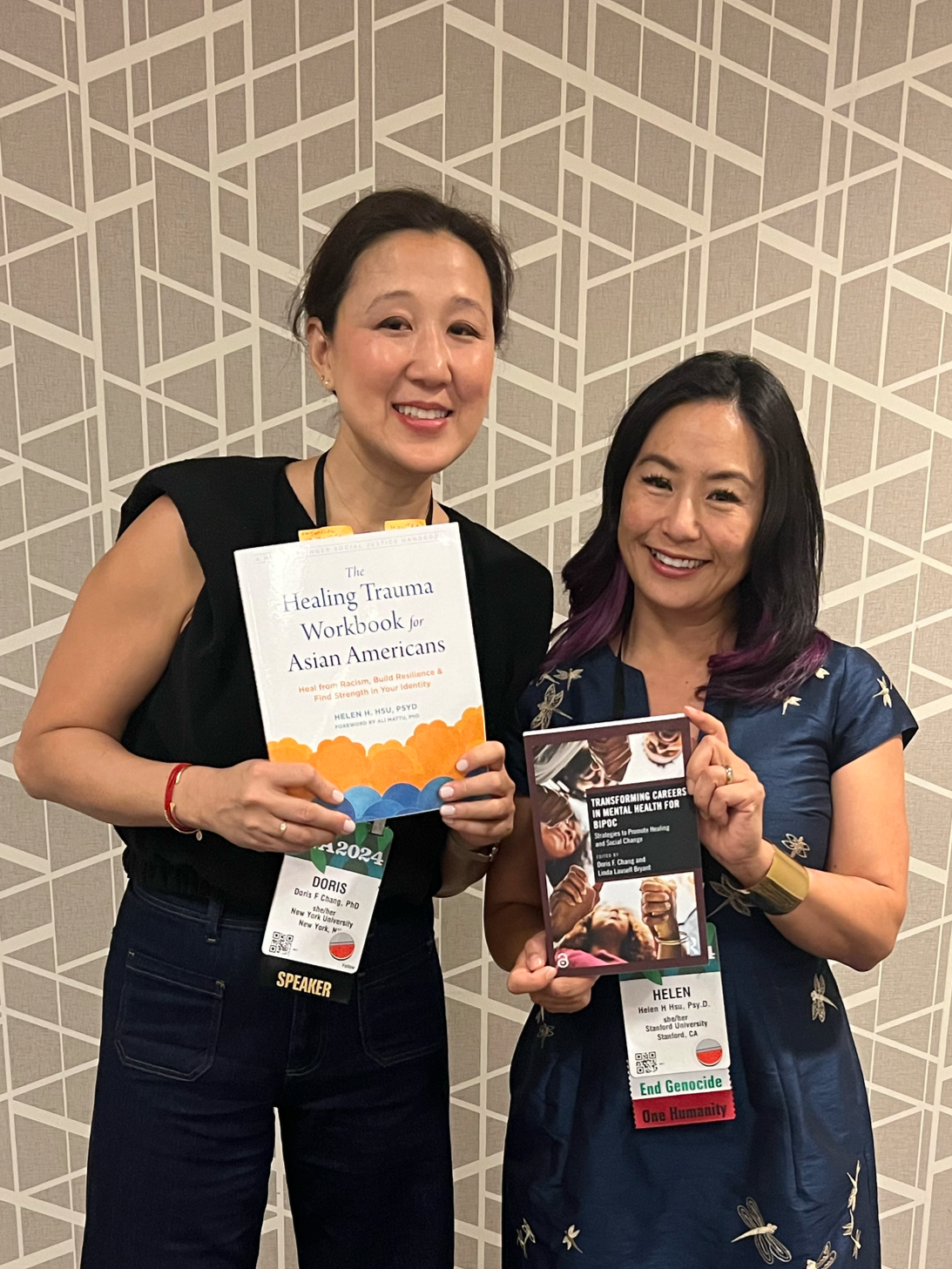 Two women standing together, each holding a book. The woman on the left has short dark hair, is smiling, and wears a black sleeveless top and dark jeans. She holds a book titled 'Healing Trauma Workbook for Asian Americans.' The woman on the right ha