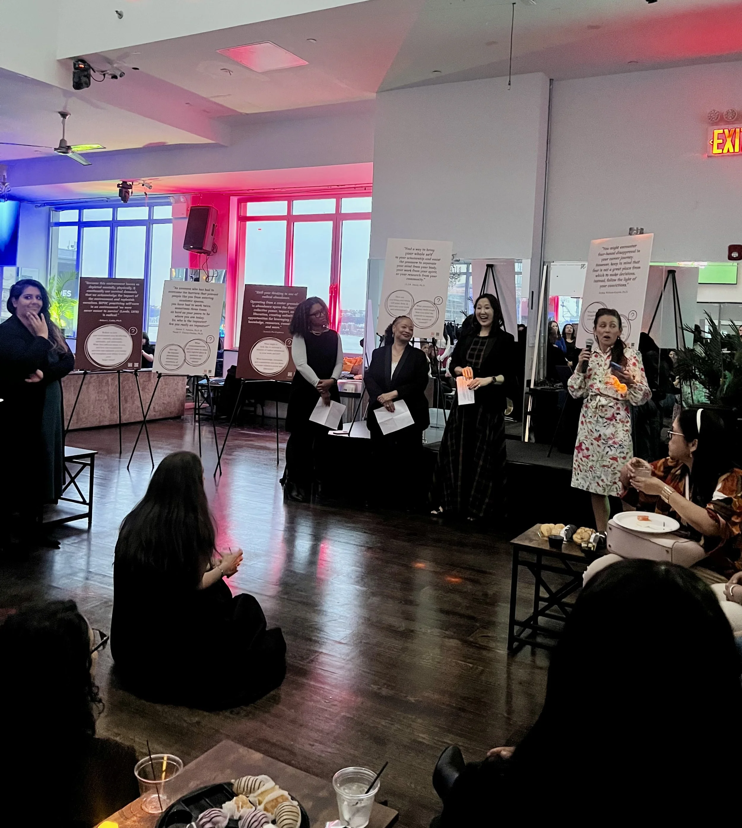 A group of women standing and sitting in a room with wooden flooring, colorful lighting, and posters on easels, during an event or presentation.