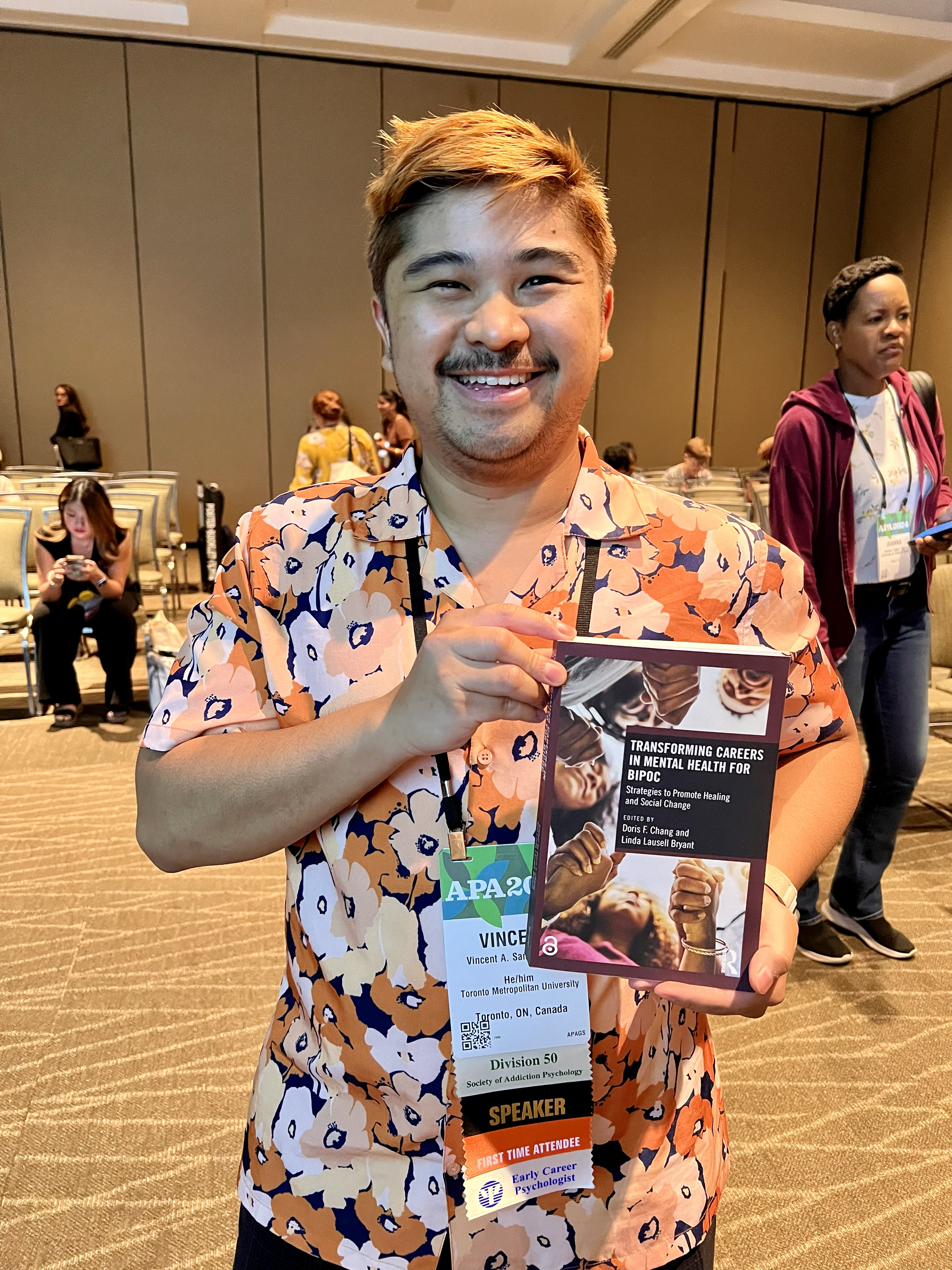 A smiling man with light brown hair and a mustache holding a book at a conference. He is wearing a floral shirt and a conference badge that says 'SPEAKER' and 'FIRST TIME ATTENDEE'.