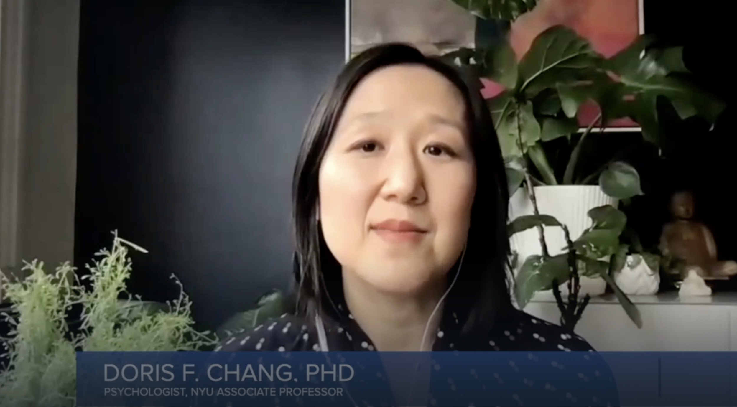 A woman with medium-length black hair, wearing a dark polka-dot top, is speaking during a virtual interview or presentation. She is sitting in front of green houseplants and artwork, with a caption overlay identifying her as Doris F. Chang, PhD, a psychologist and associate professor at NYU.