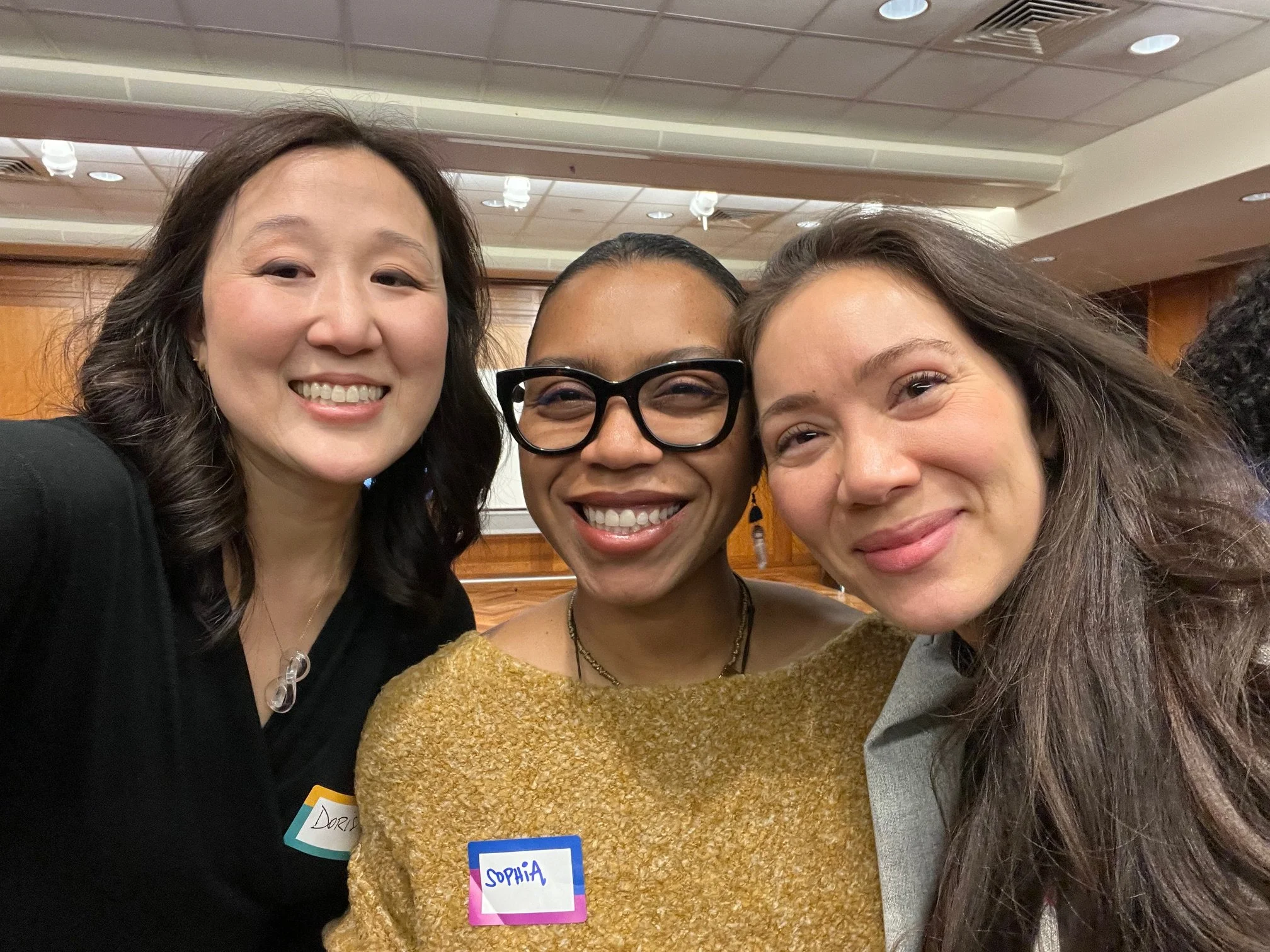 Three women smiling and taking a close-up selfie together at an indoor event. They are wearing name tags, and one of the women has glasses. The background shows a ceiling with lights and wood paneling.