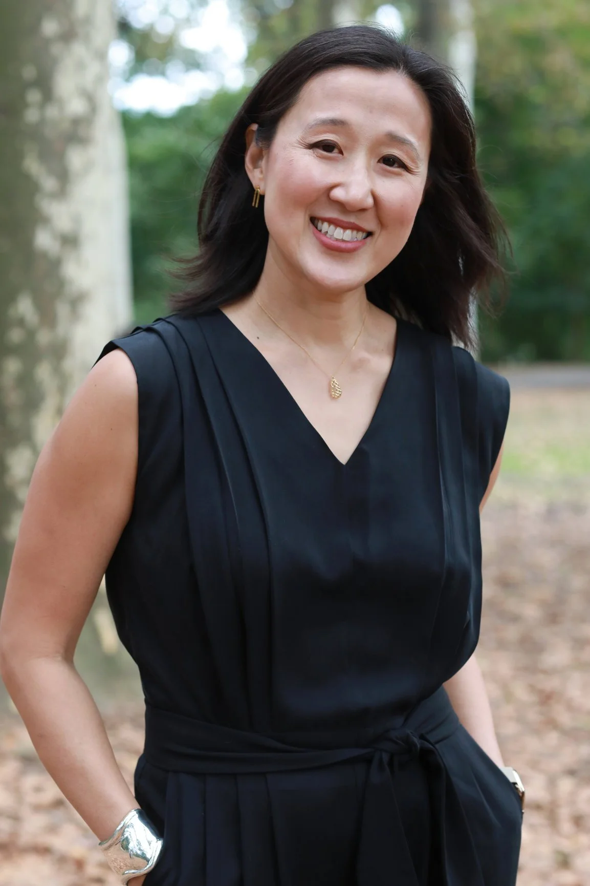 A smiling woman in a black sleeveless dress outdoors, standing near trees with green foliage.
