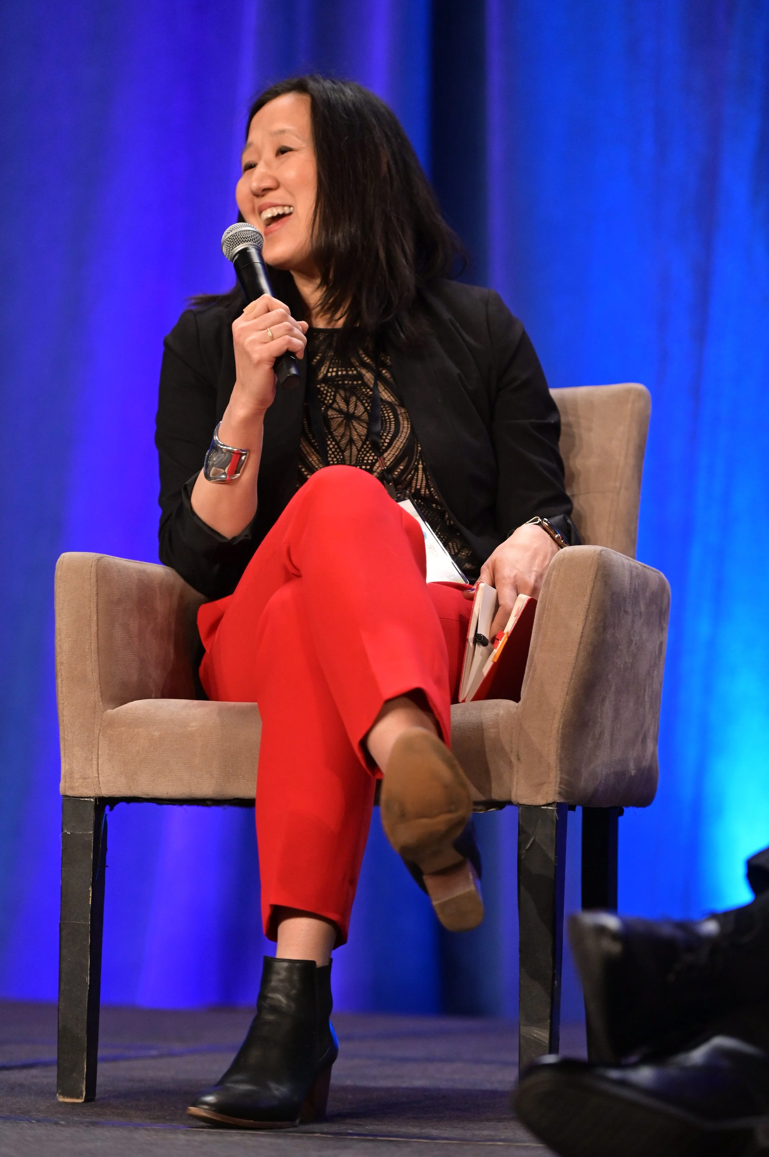 A woman with black hair, wearing a black blazer, red pants, and black ankle boots, sitting on a beige chair, holding a microphone, and speaking at an event with a blue backdrop.