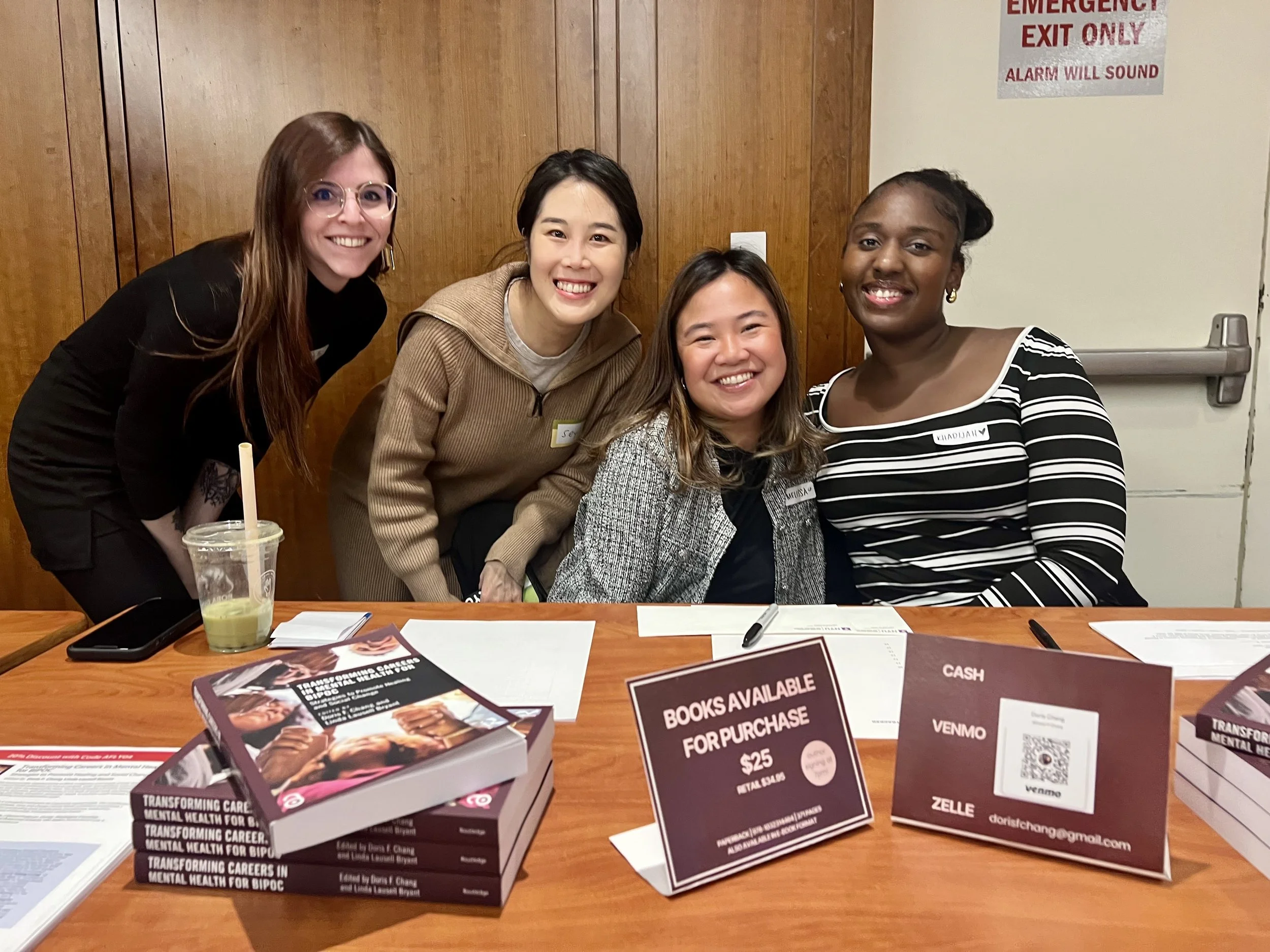 Four women smiling and gathered together behind a table at a book signing event. The table has stacks of books, pamphlets, and signs displayed, with one sign indicating books are available for purchase for $25. The background includes a wooden wall a
