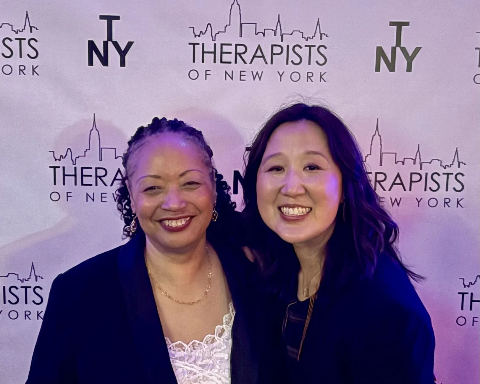 Two smiling women standing together in front of a backdrop with the text 'Therapists of New York' and a city skyline outline. One woman has curly hair and is wearing a black blazer and earrings; the other has straight dark hair and is wearing a dark 