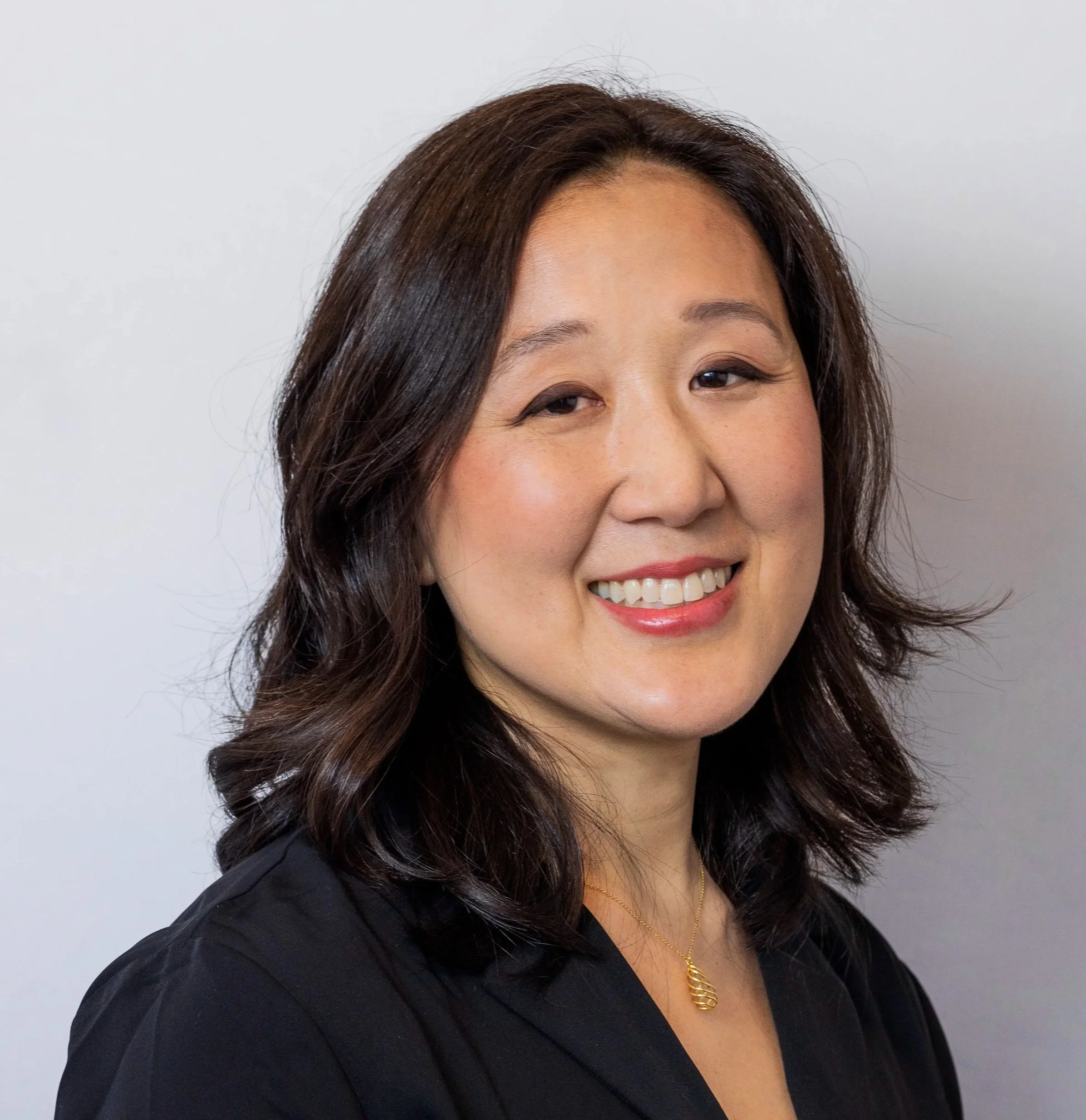 A woman with shoulder-length dark brown hair and light makeup, wearing a black top and a gold necklace, smiling against a plain white background.